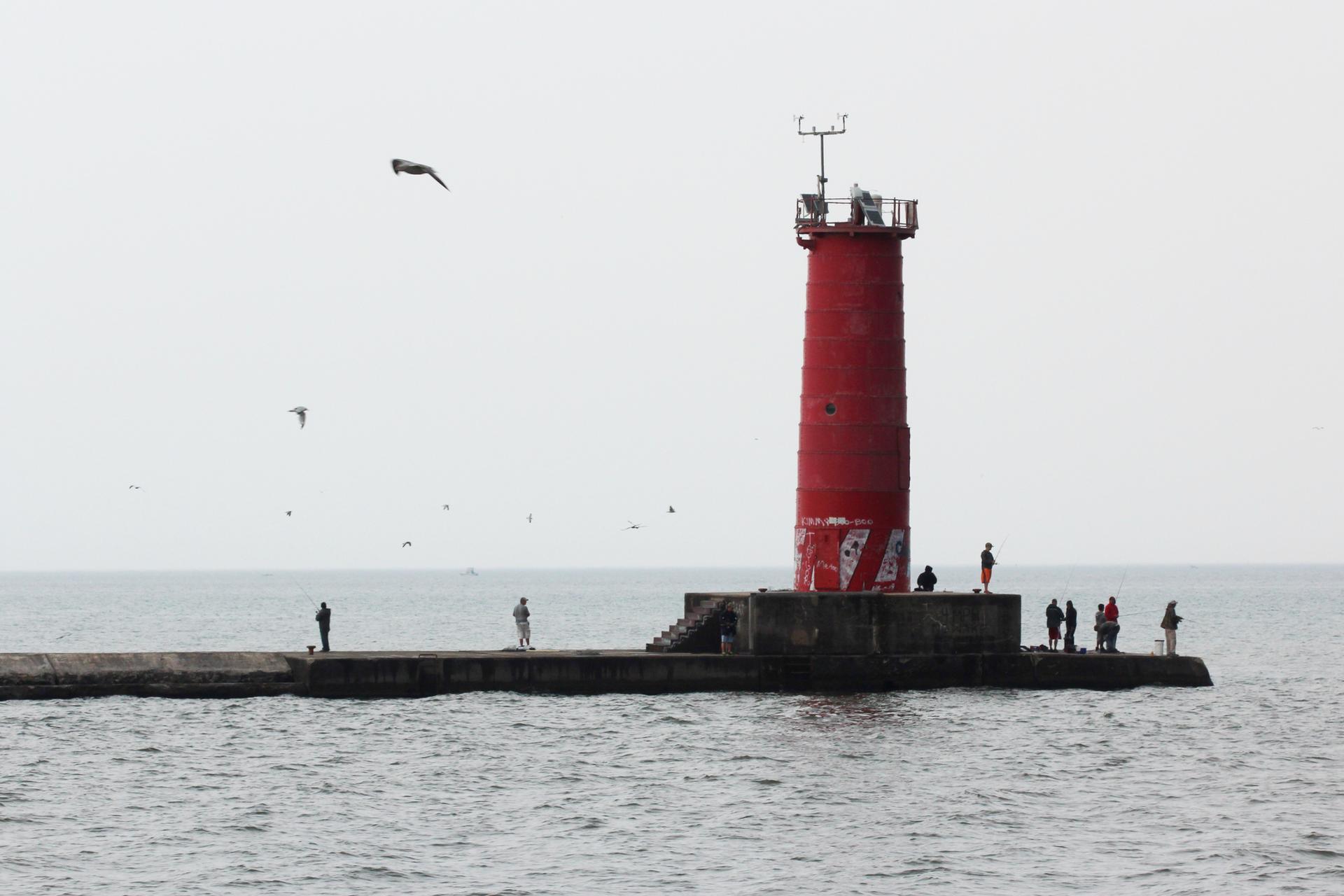 Red lighthouse on a pier with people standing and birds flying overhead.