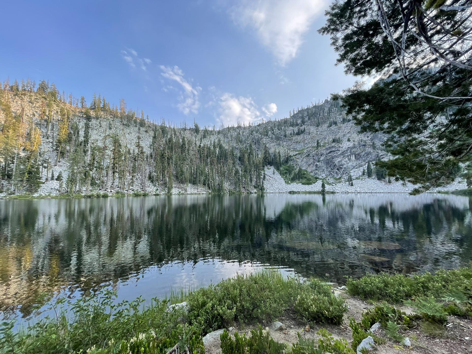 Calm lake reflecting forested hills under a bright blue sky.