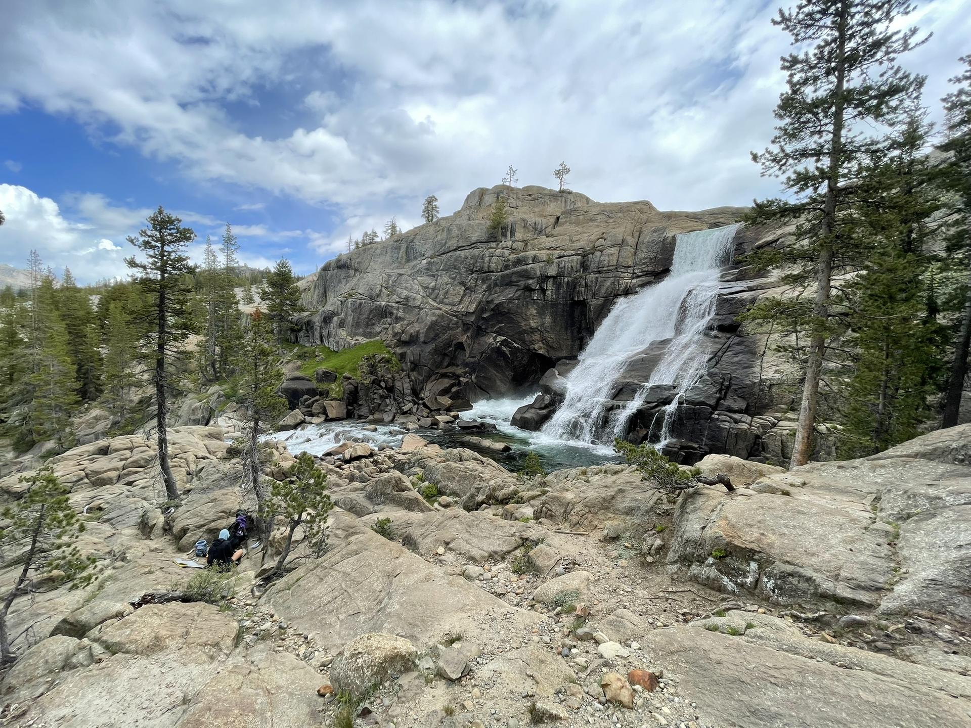 A large waterfall tumbles over granite rocks into a pool, surrounded by trees.
