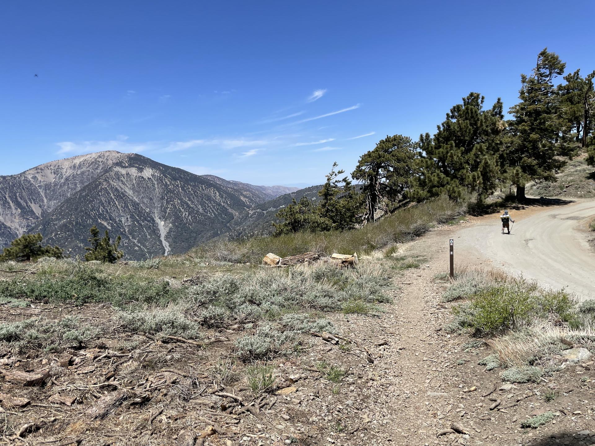 Person walks on a dirt road overlooking vast mountains under a clear blue sky.