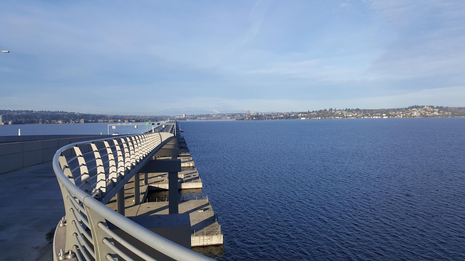 Curved pedestrian bridge over a wide, calm lake under blue sky.