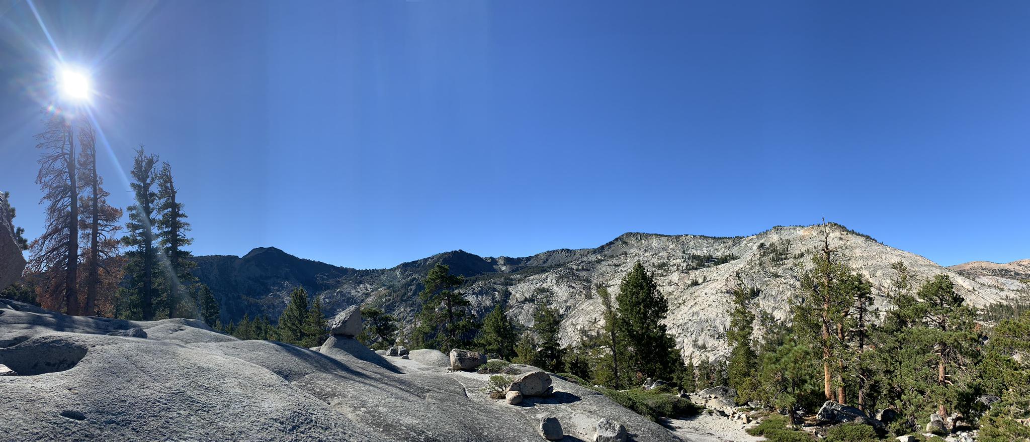 Bright sun over a rocky landscape with pine trees and distant mountains.