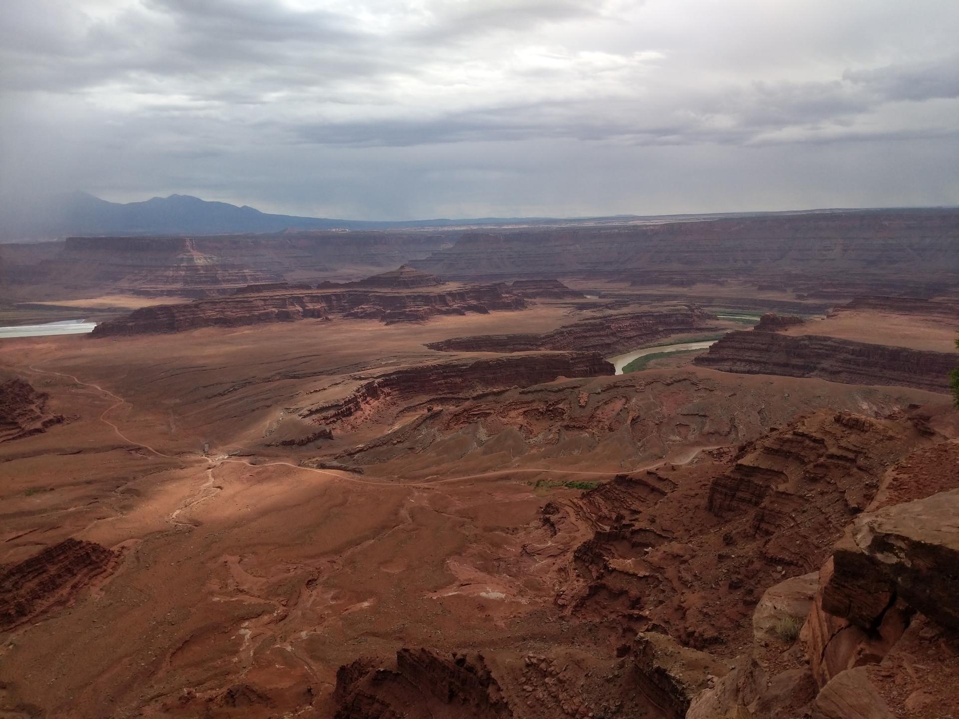 Broad desert landscape with canyons and a distant river under a cloudy sky.
