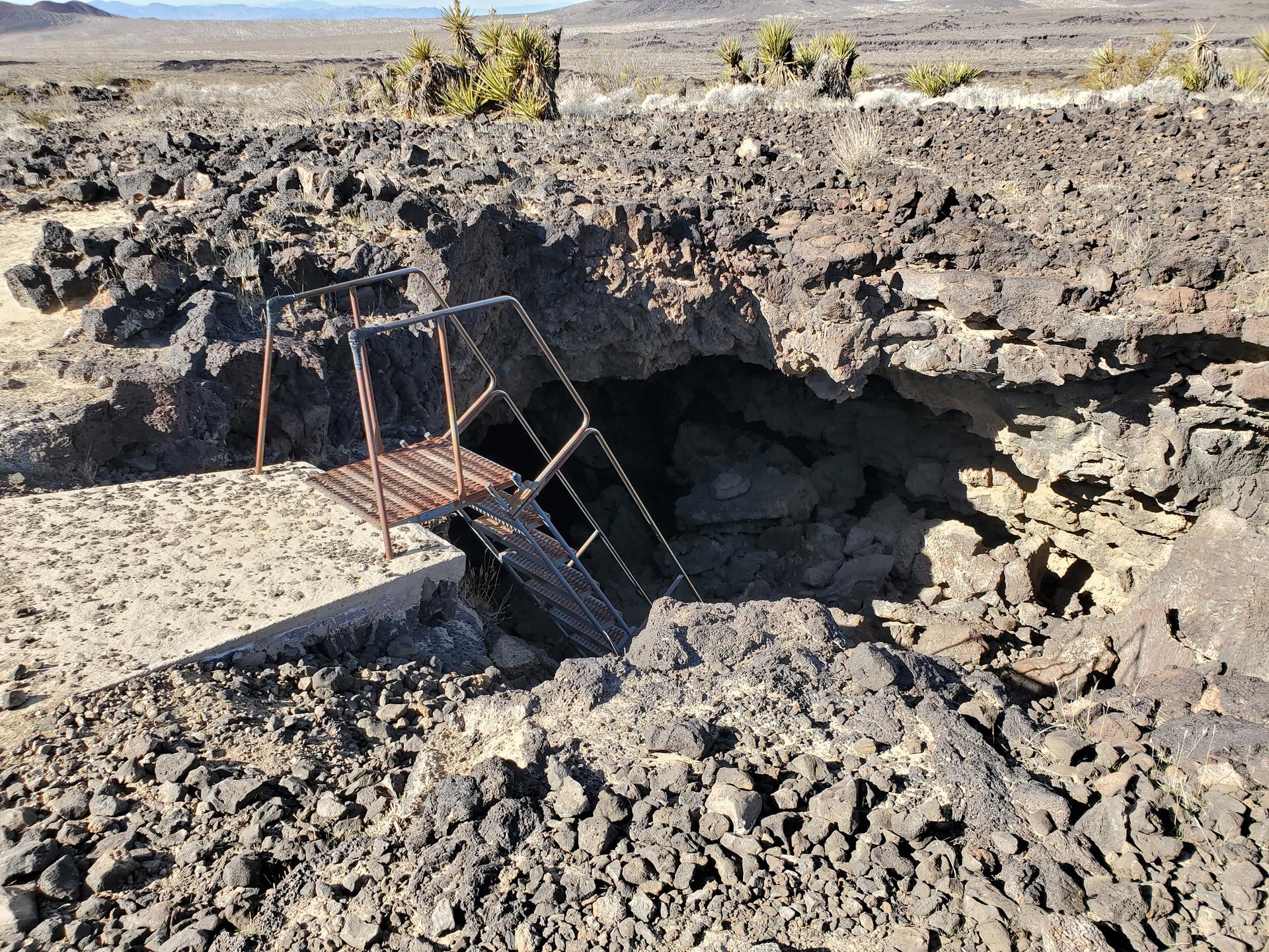Metal stairs lead into a dark cave entrance in rocky desert terrain.