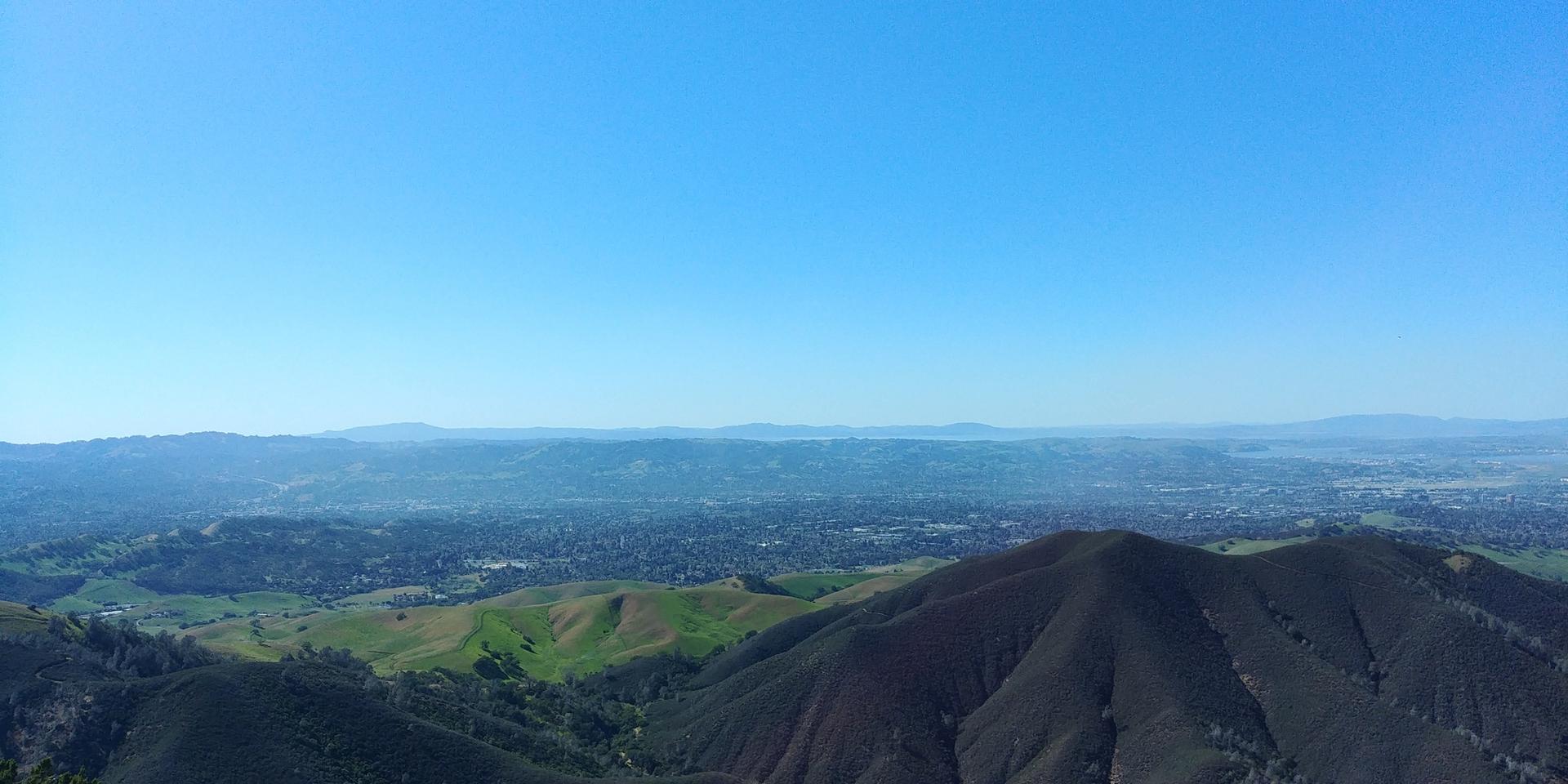 Vast panoramic view of green and dark mountains, with a distant city. Clear sky.