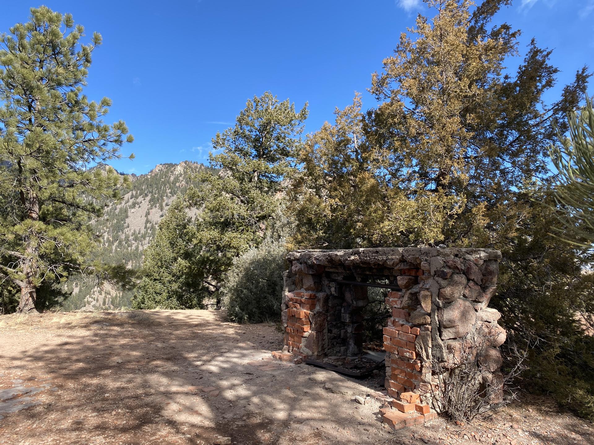 Stone structure in a clearing, surrounded by pine trees with mountains in background.
