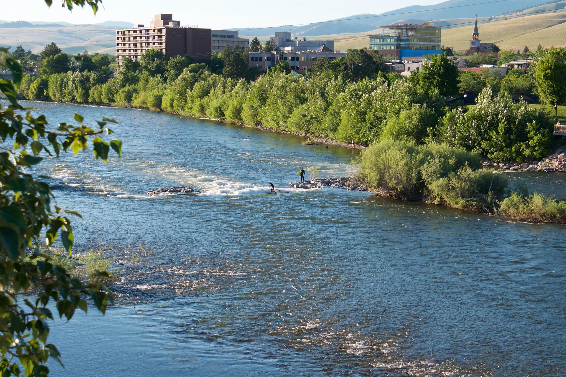 Two people paddleboarding on a wide river with a city and hills in the background.