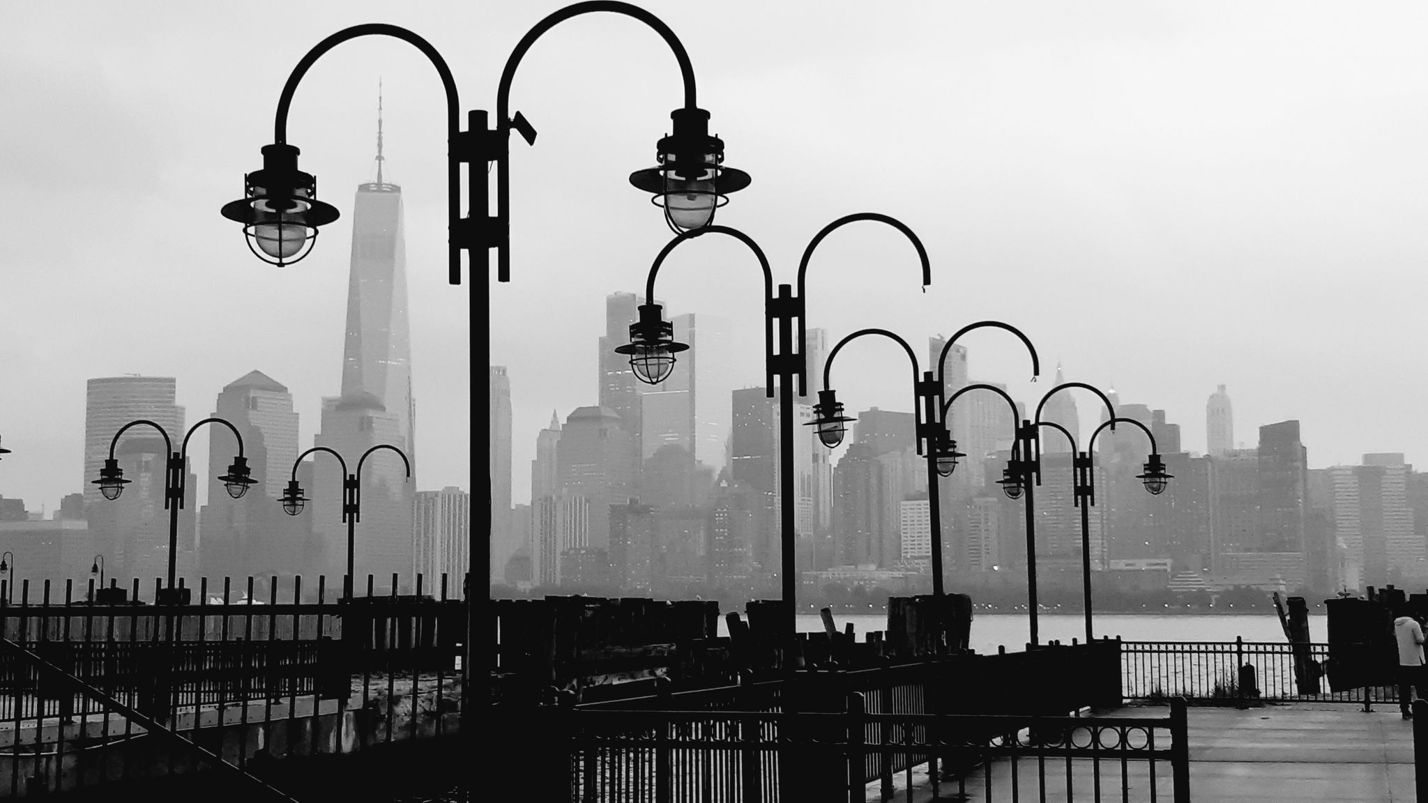 Silhouetted streetlights against a hazy city skyline, in black and white. - Hudson River Waterfront Walkway