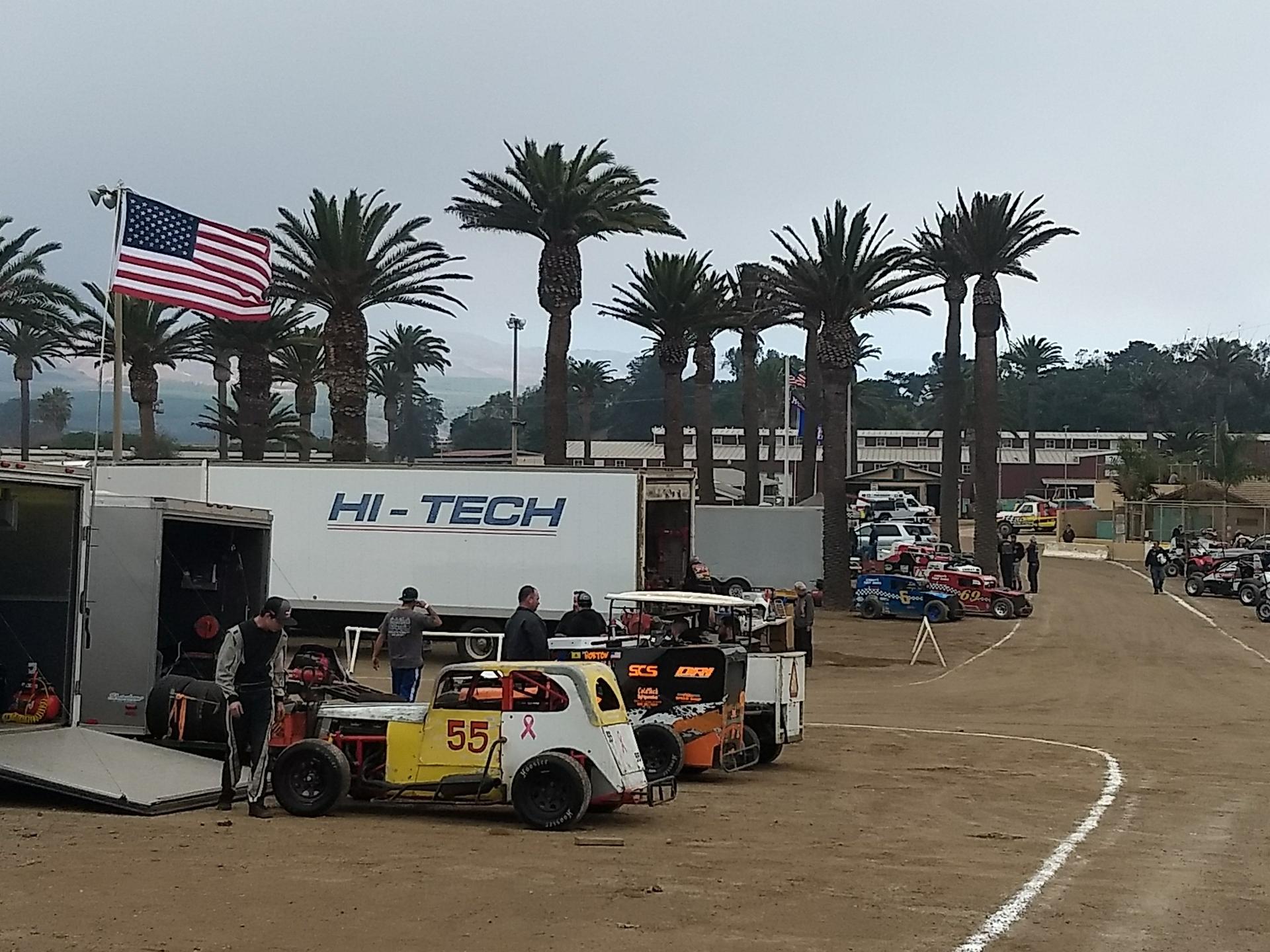 People and race cars gathered on a dirt track under palm trees, overcast day.