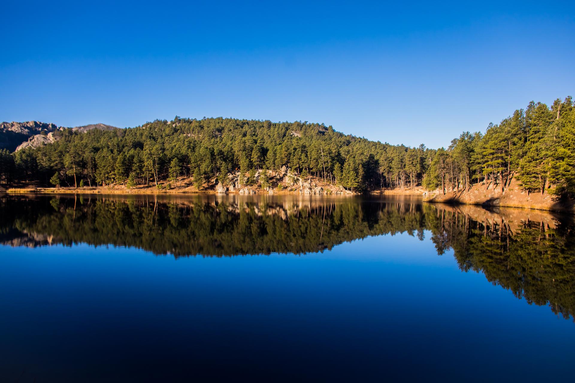 Calm lake reflecting forested hills under a clear blue sky.