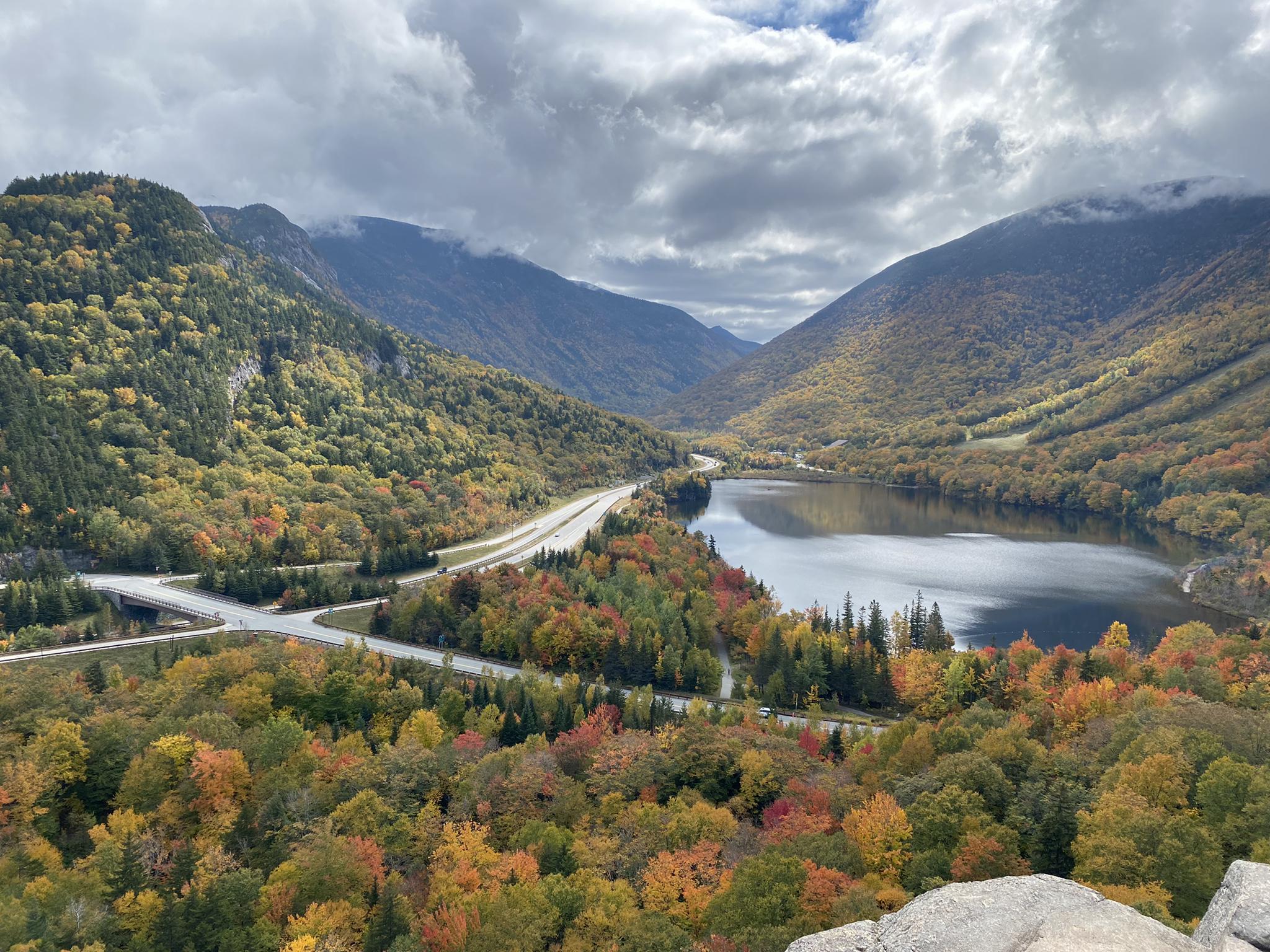 Lake and highway winding through mountains with vibrant autumn trees, cloudy sky.