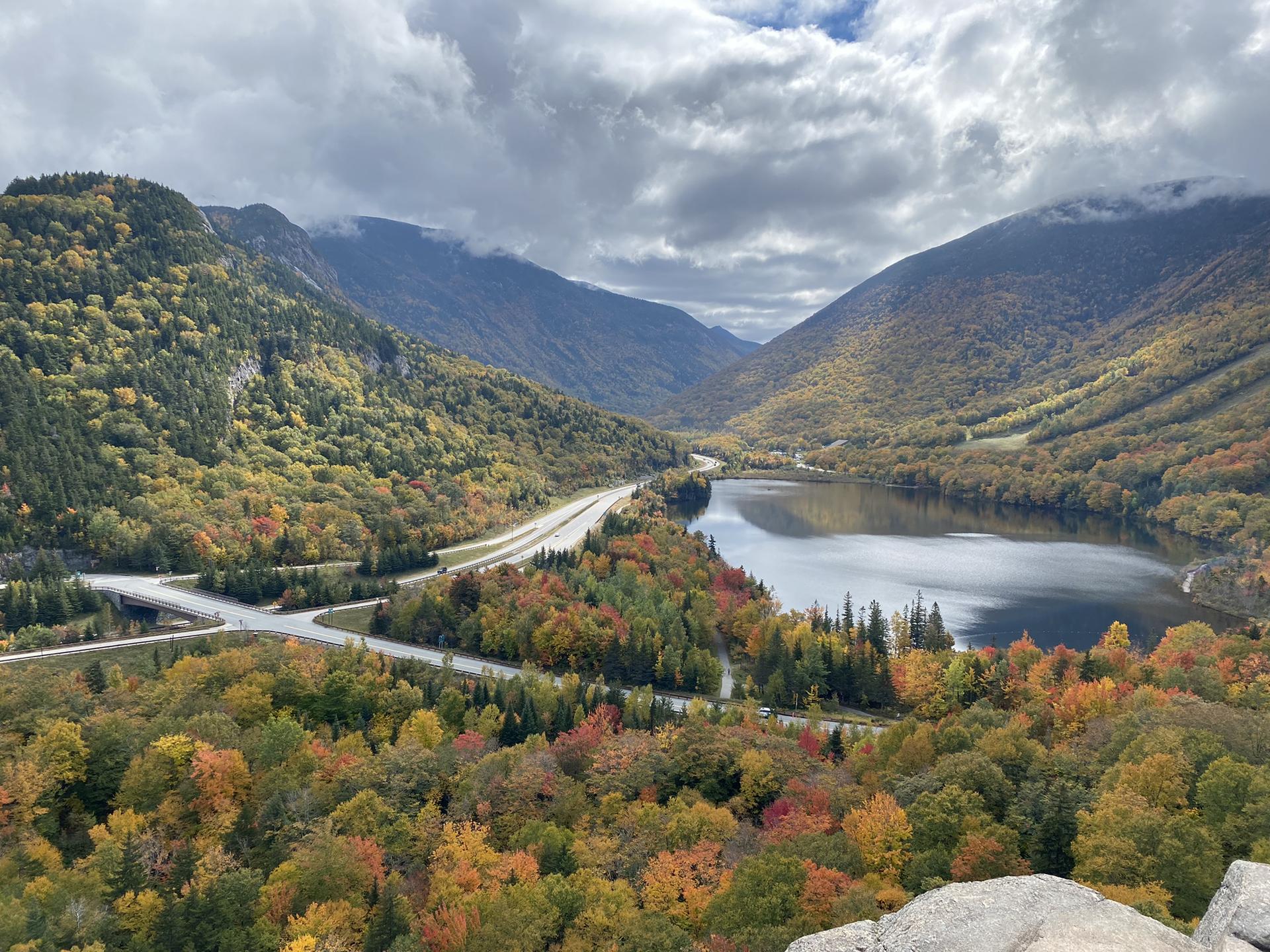 Lake and highway winding through mountains with vibrant autumn trees, cloudy sky.