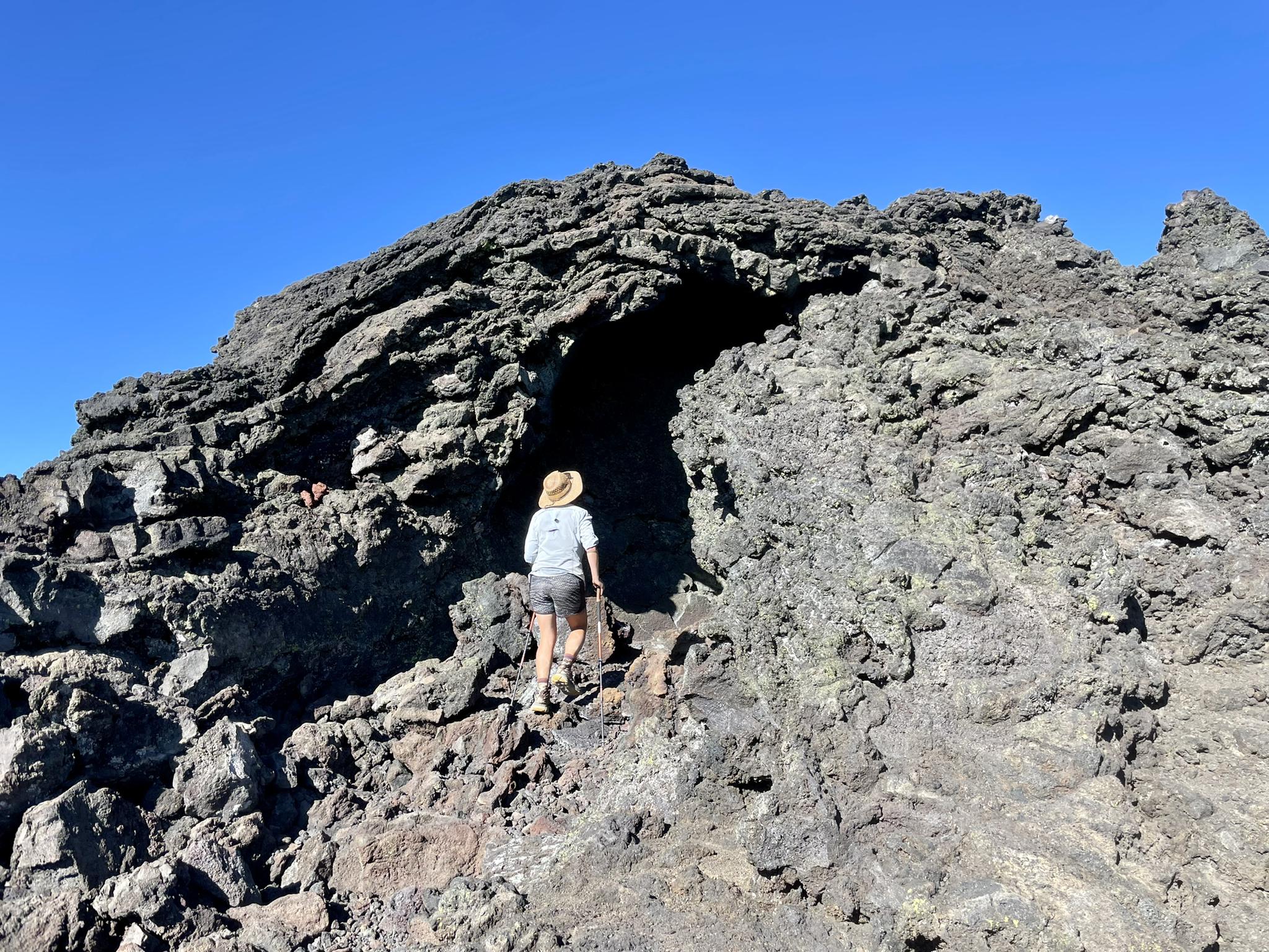 A person in a hat climbs into a dark, rocky lava cave entrance.