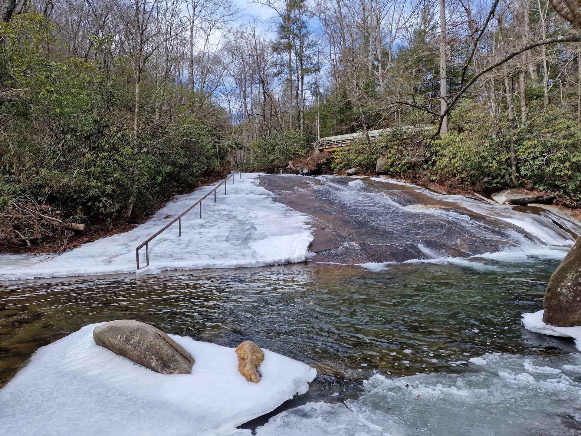 Icy river flows over a wide rock slide with metal handrails.