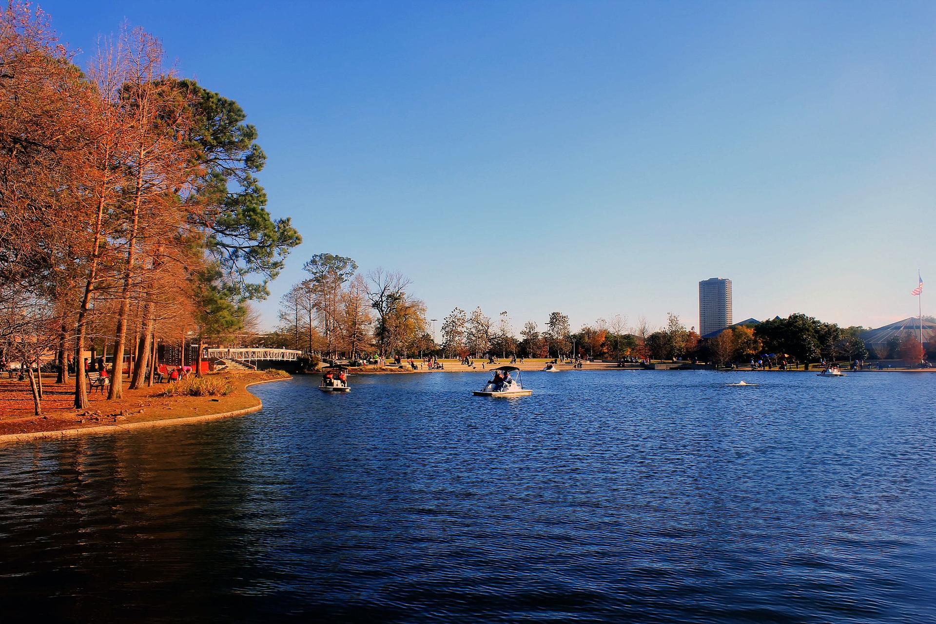 People paddle boating on a lake surrounded by autumn trees and a distant building.