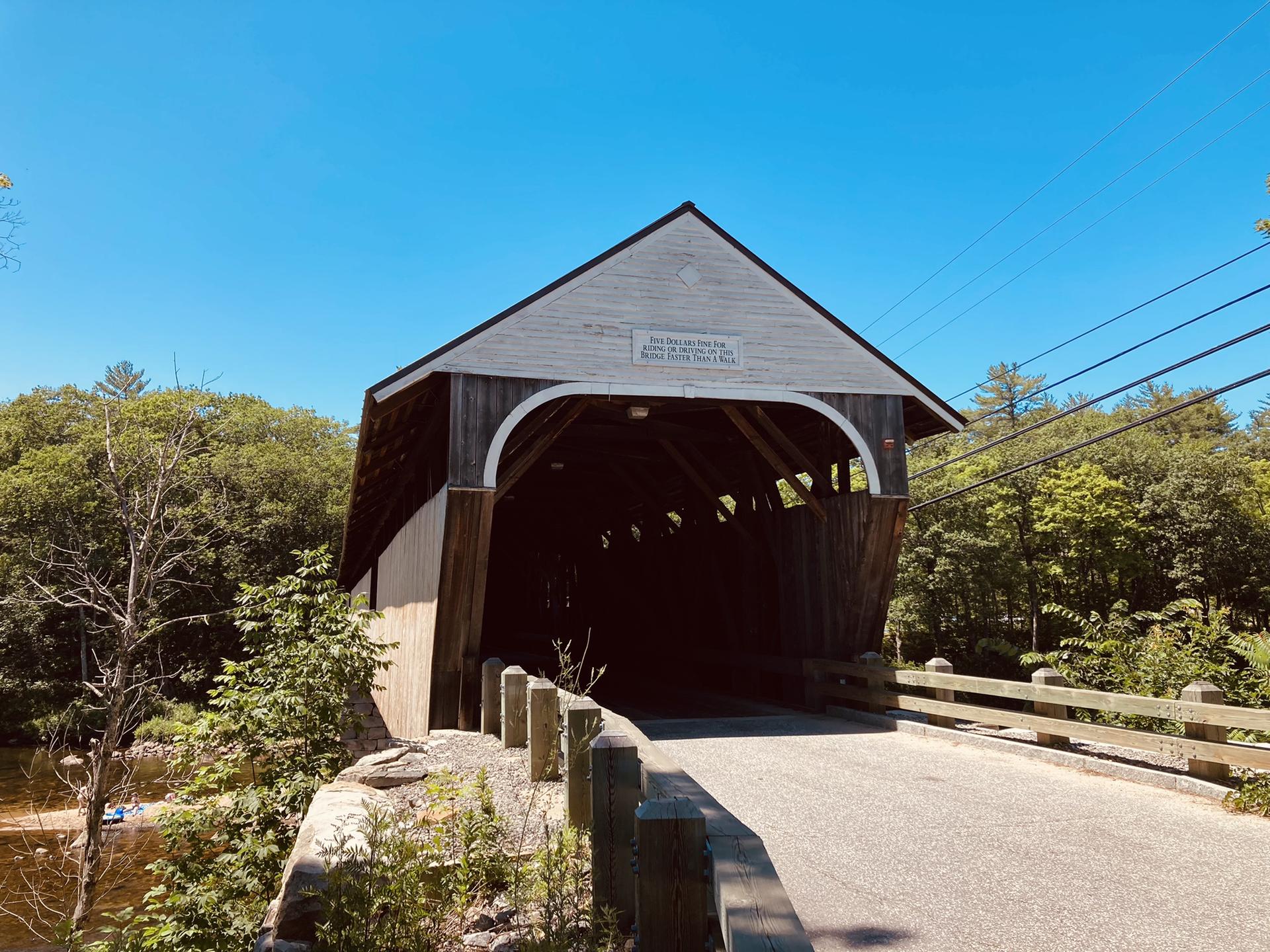 A rustic wooden covered bridge spans a river on a bright day.