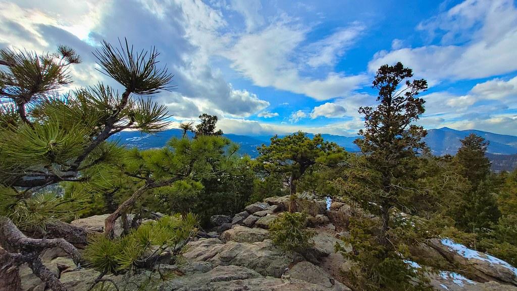 Pine trees and rocky terrain overlooking distant mountains under a cloudy sky.
