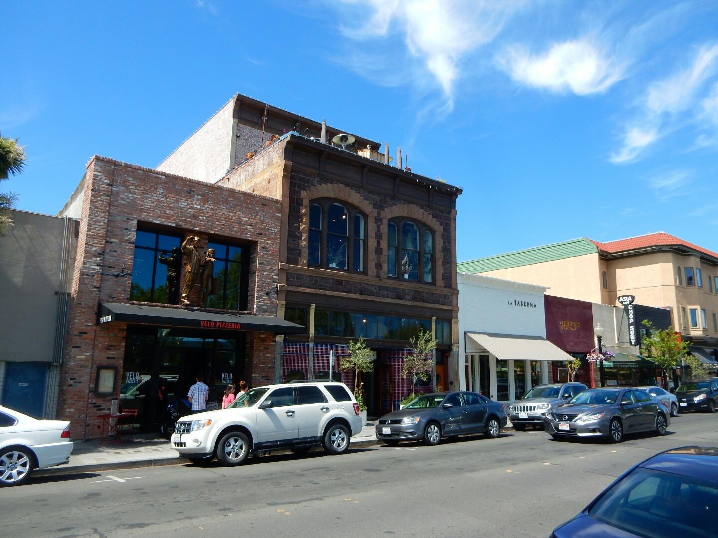 Sunny urban street with historic brick buildings, parked cars, and people.