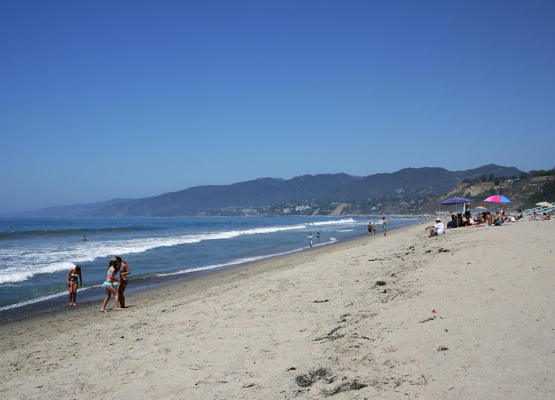 Sunny beach scene with people walking by the ocean and distant mountains.