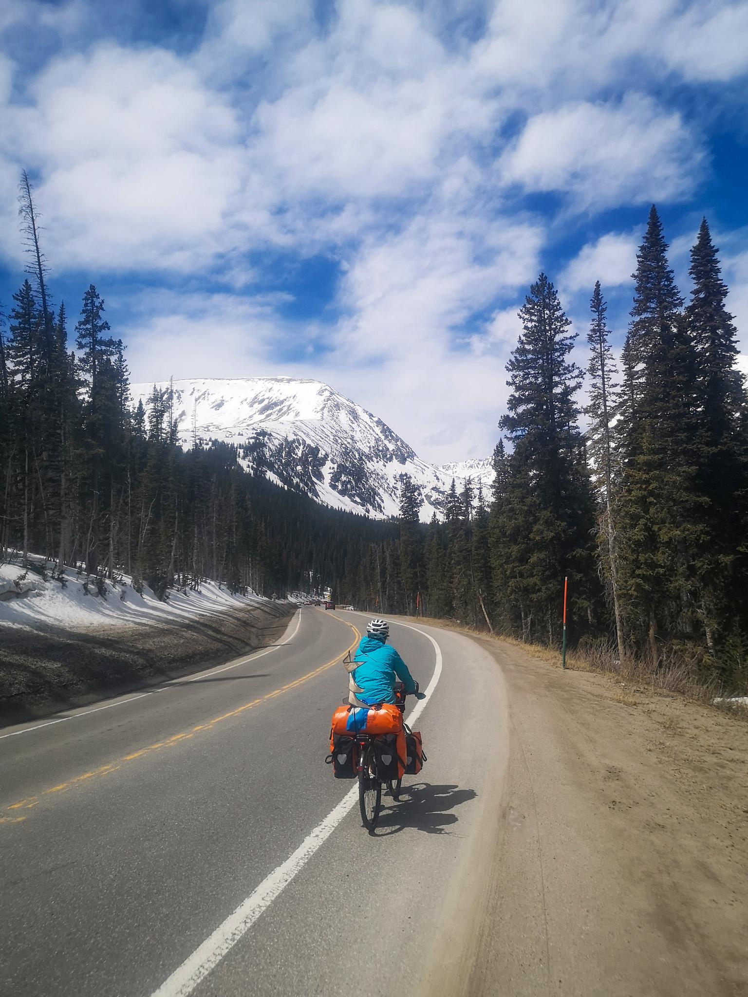 A cyclist rides a paved mountain road towards a snow-capped peak.