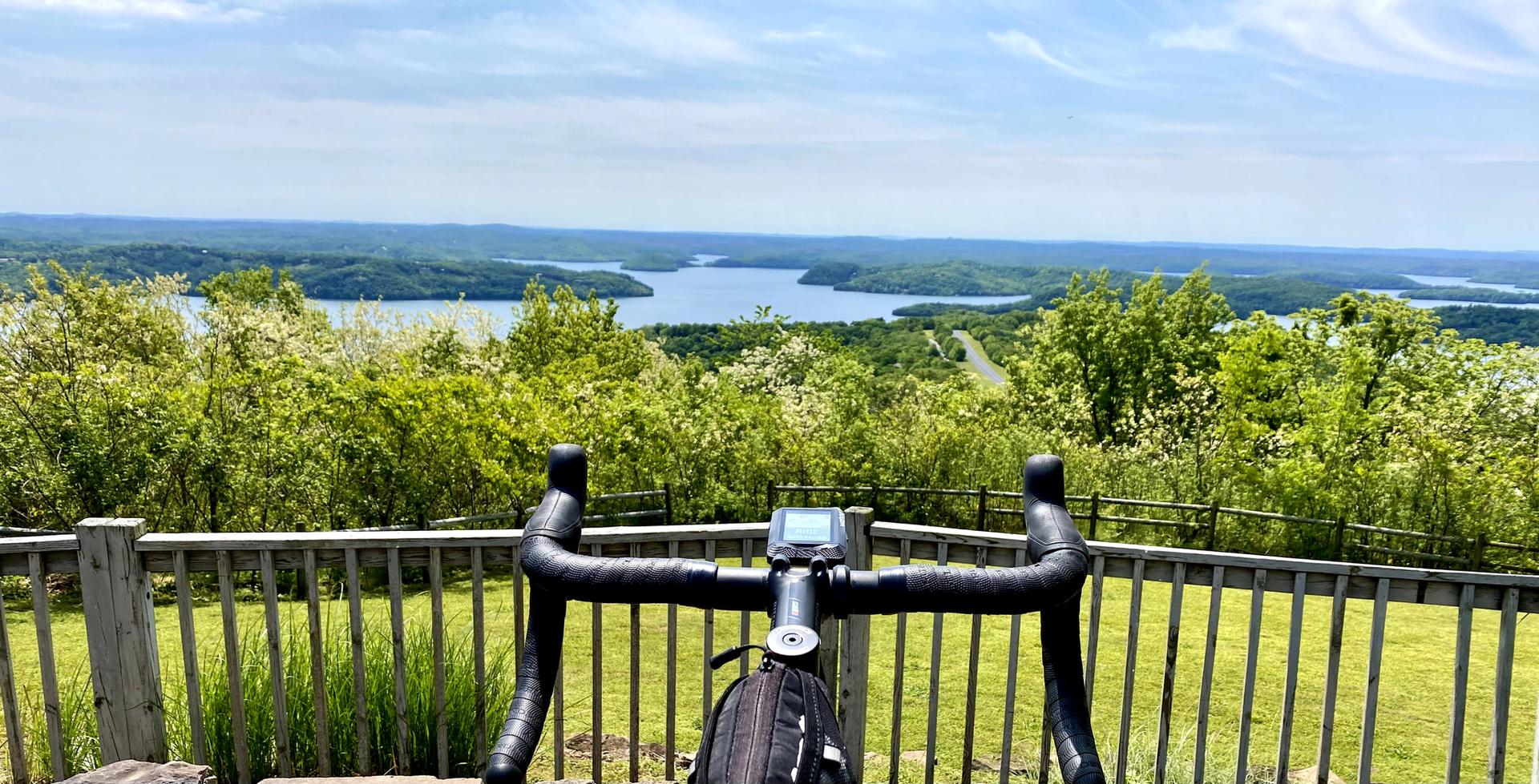 Bicycle handlebars with a scenic view of a lake and green forested hills.
