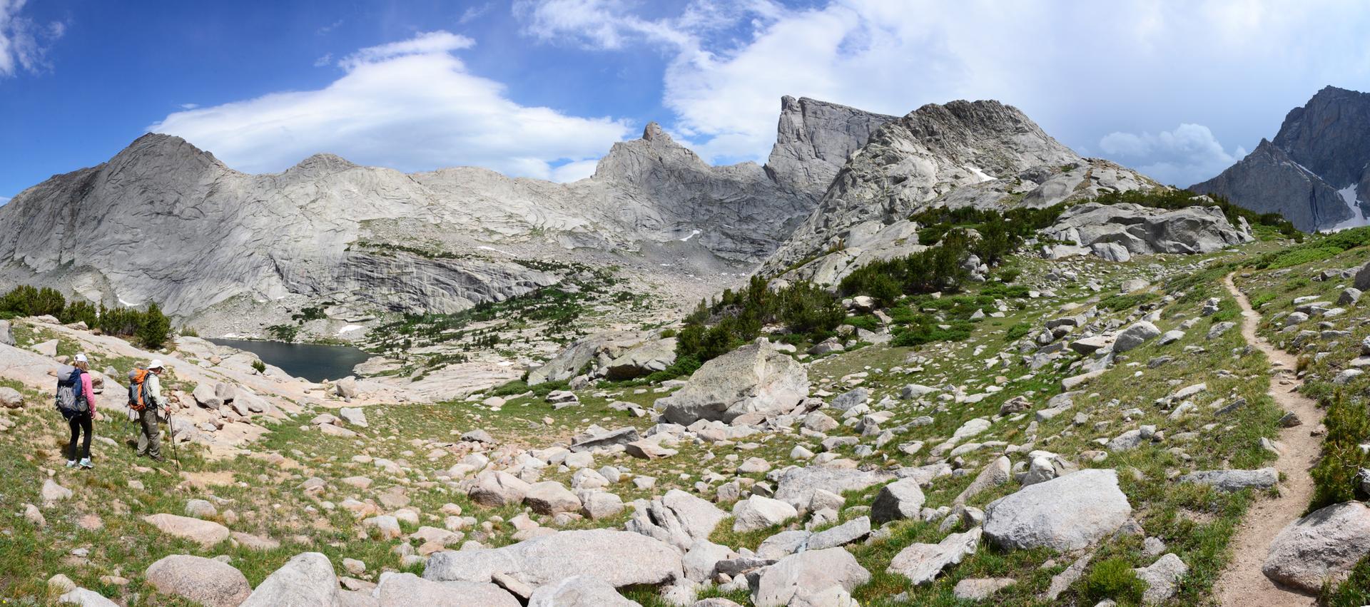 Two hikers on a rocky trail overlooking a lake and vast mountain range.