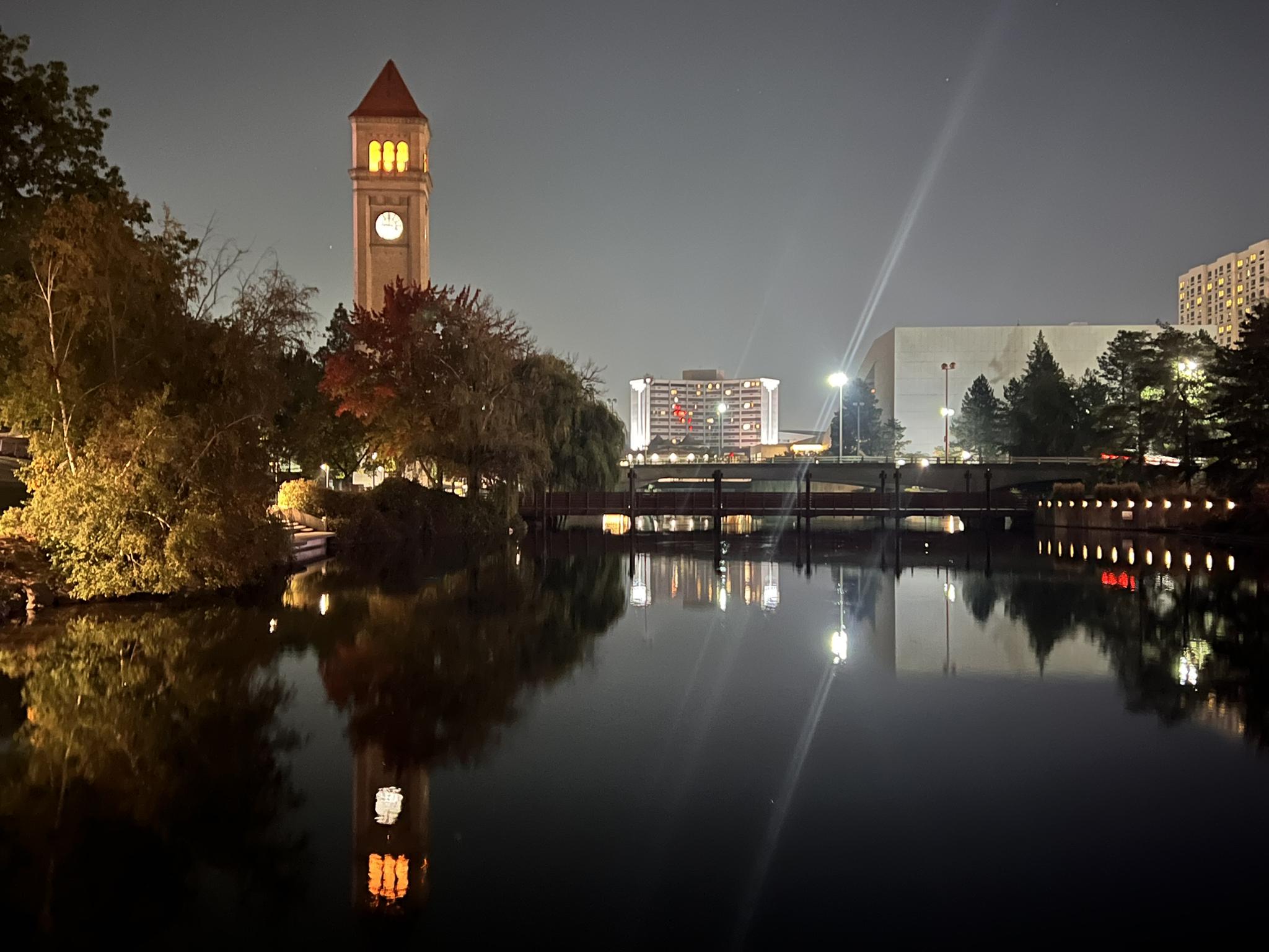 Illuminated clock tower and city buildings reflected in a dark river at night.