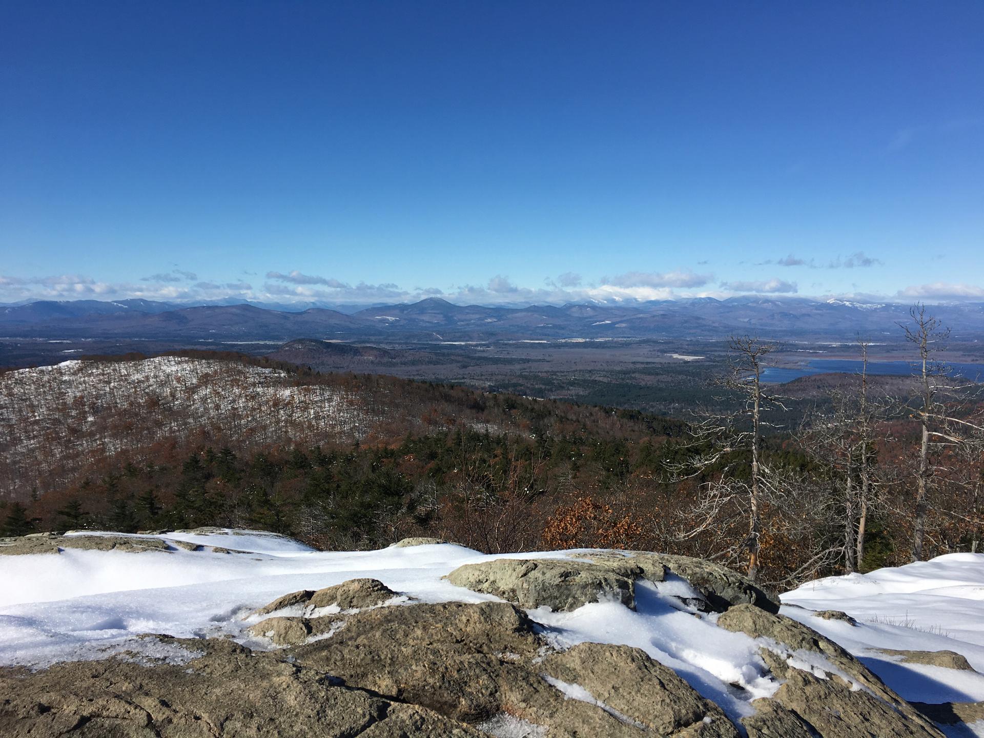 Sunny winter mountain vista with snow-covered rocks and distant peaks under a blue sky.