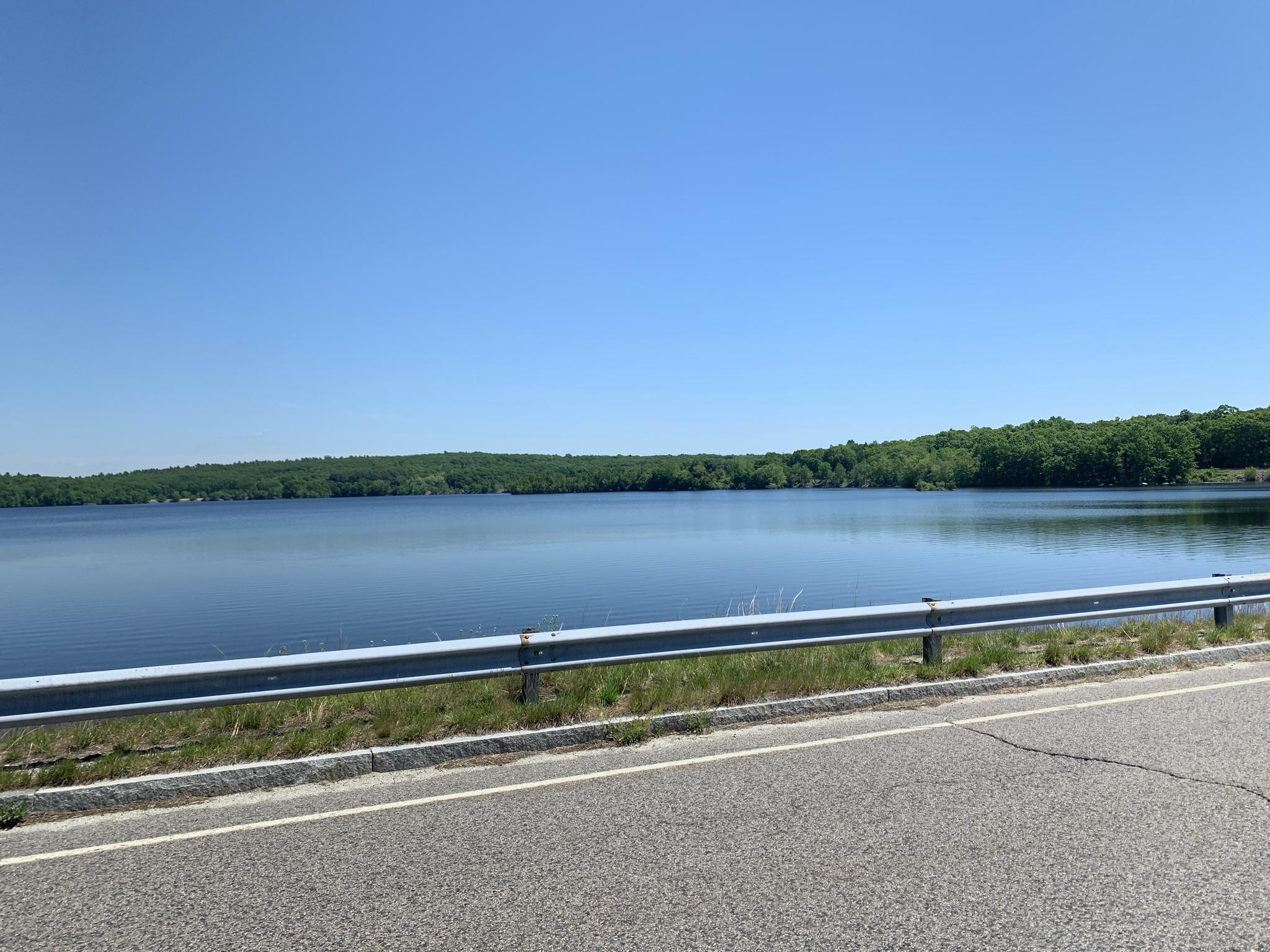 A calm lake viewed from a road with a guardrail under a clear blue sky.