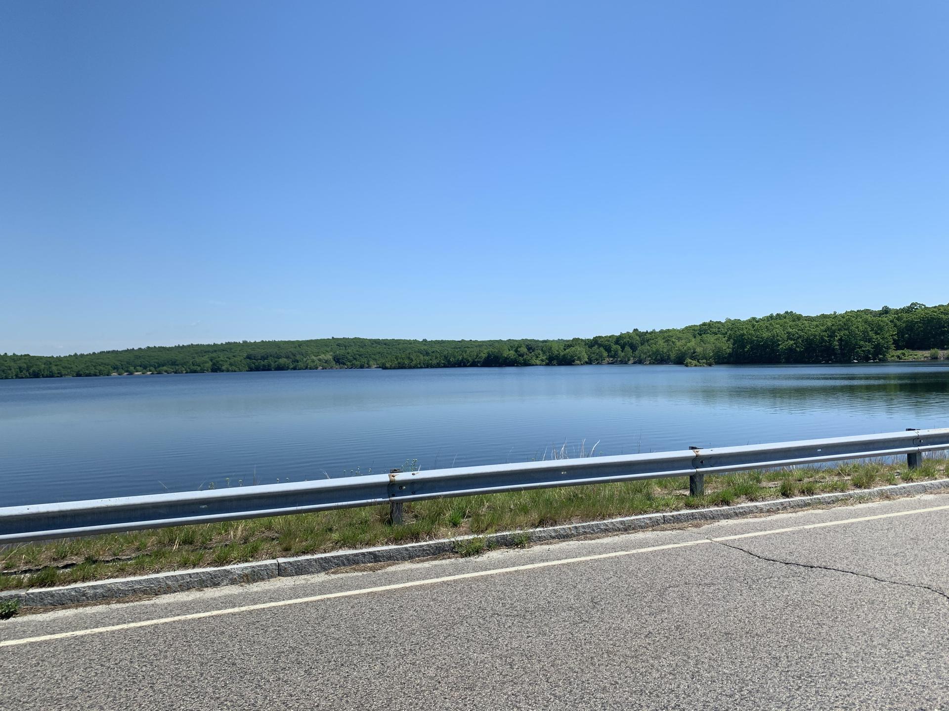 A calm lake viewed from a road with a guardrail under a clear blue sky.