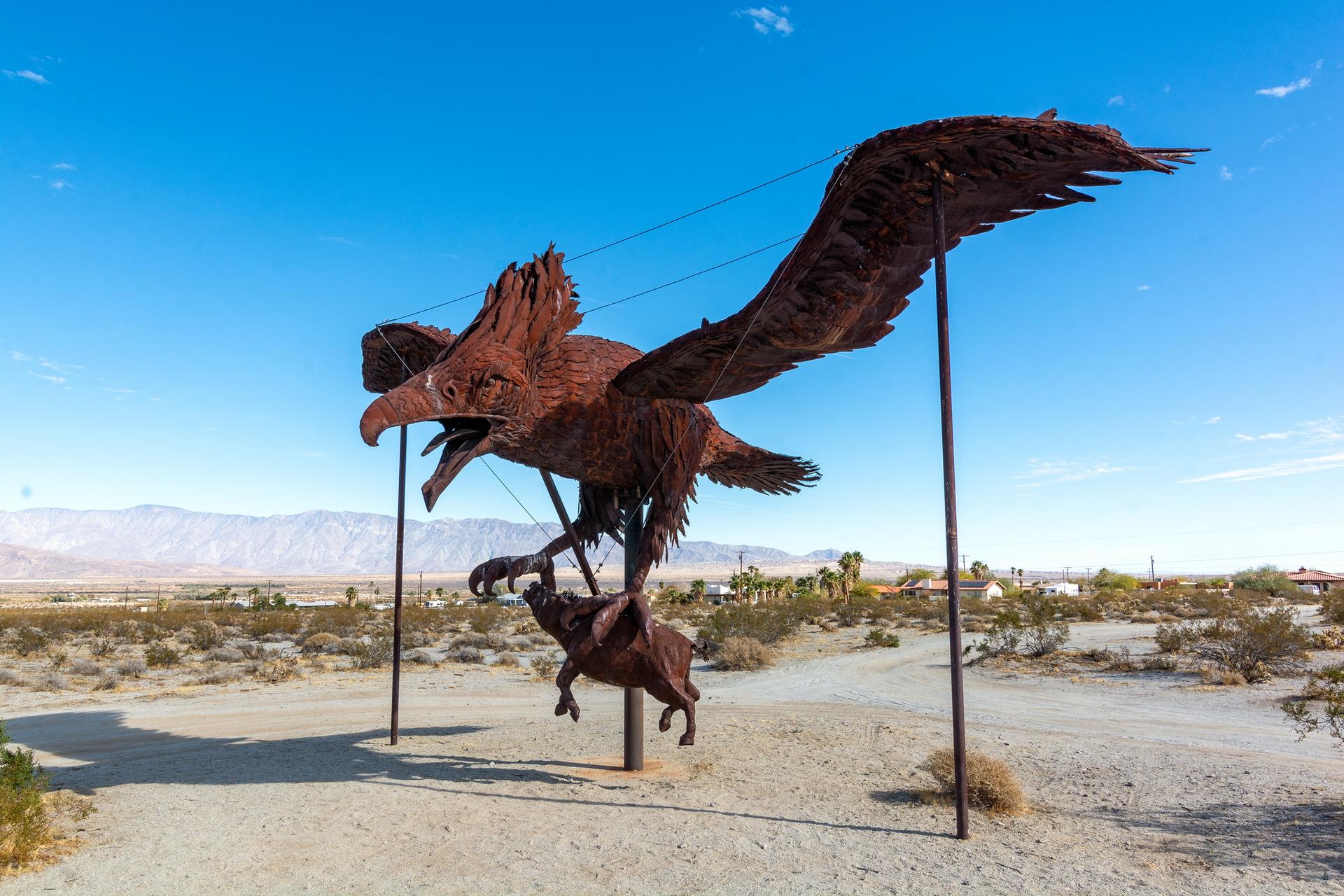 Giant metal eagle sculpture carrying a ram in a bright desert landscape.