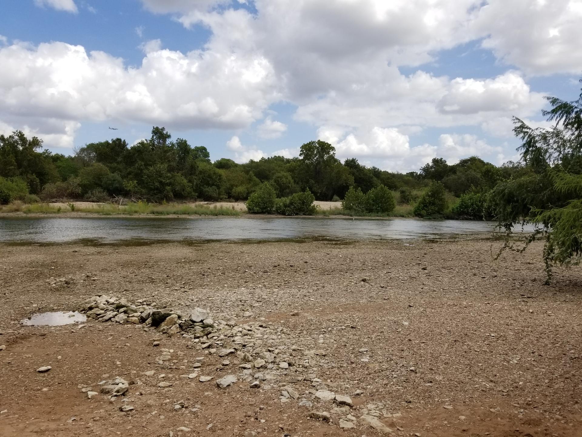 River flowing past a wide, rocky bank with green trees under a cloudy sky.