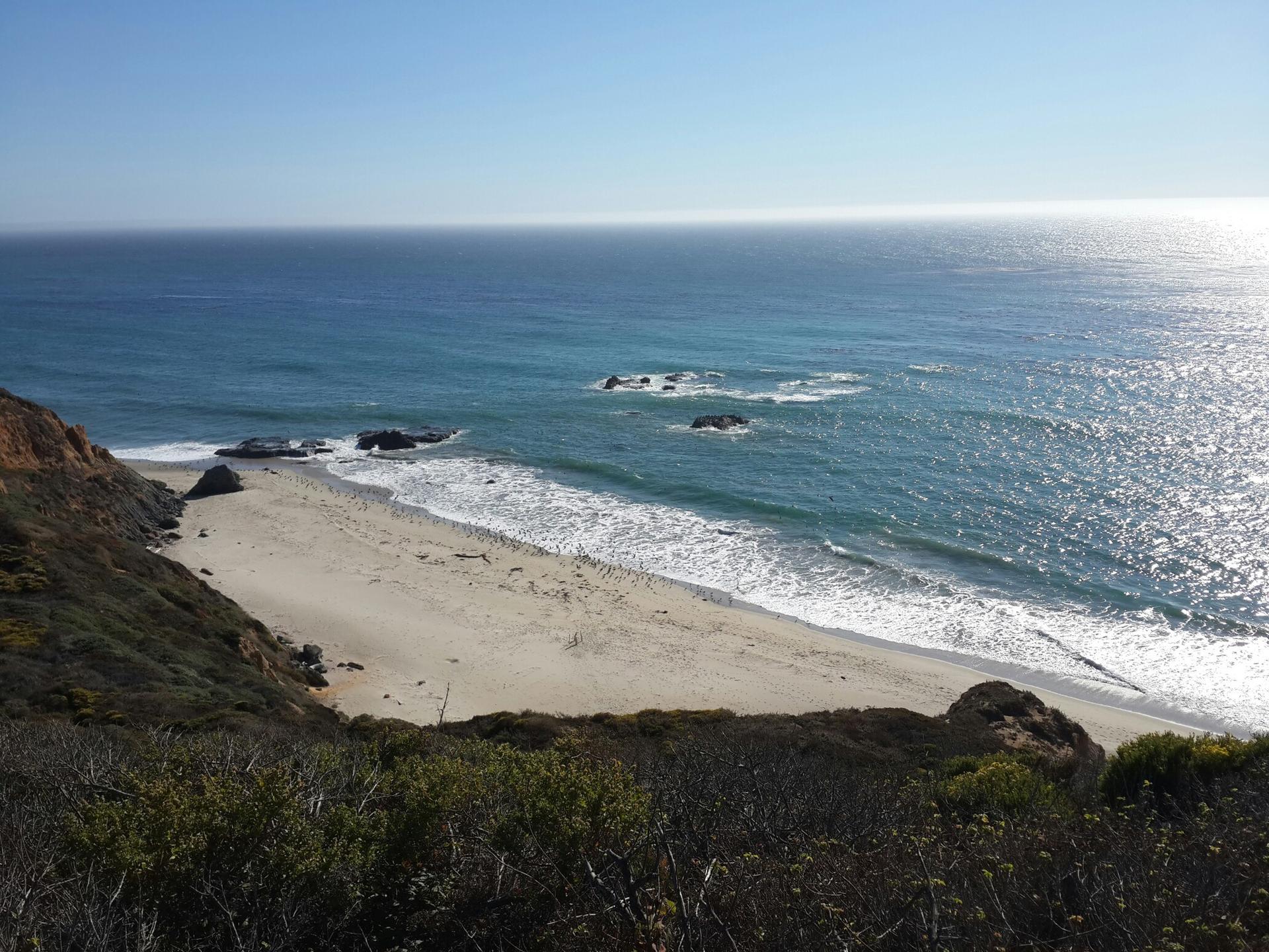 Sunny coastal view of a sandy beach, blue ocean, and green cliffs.