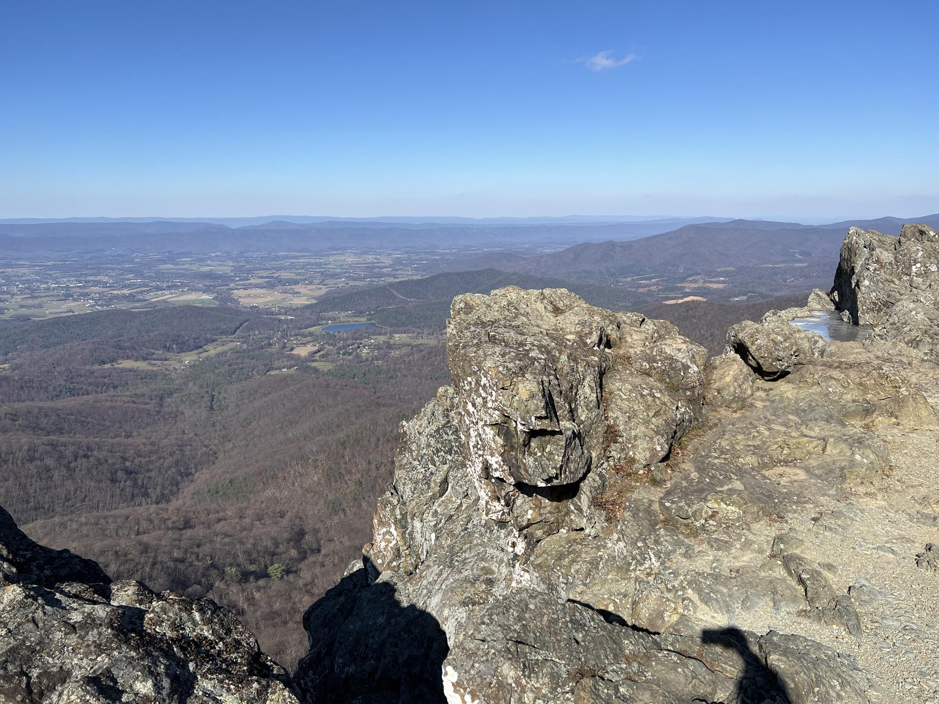 Rocky outcrop overlooks a wide, hazy valley with sparse trees under clear sky.