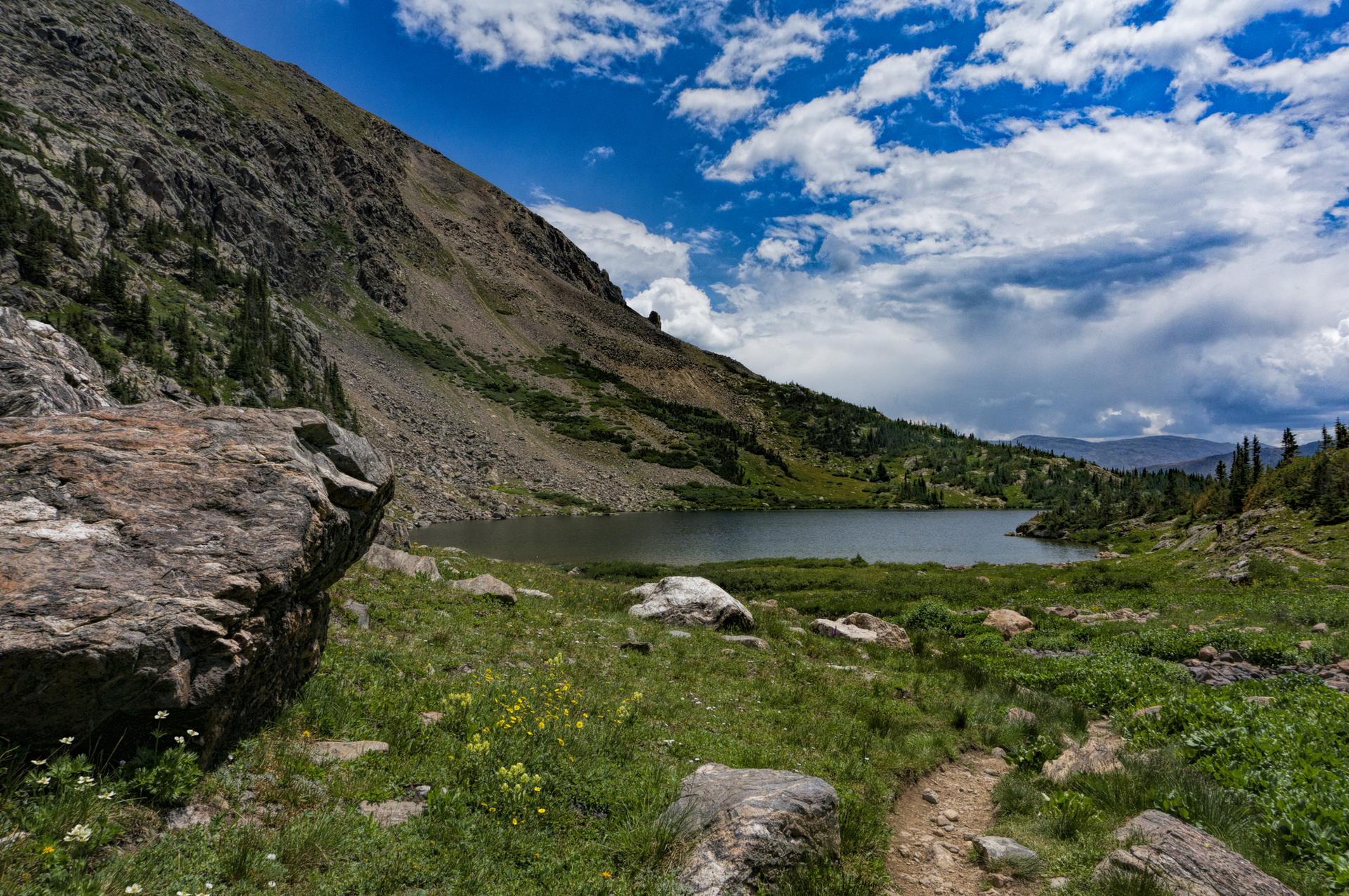 Mountain lake with a rocky shore, green grass, and a dirt path.