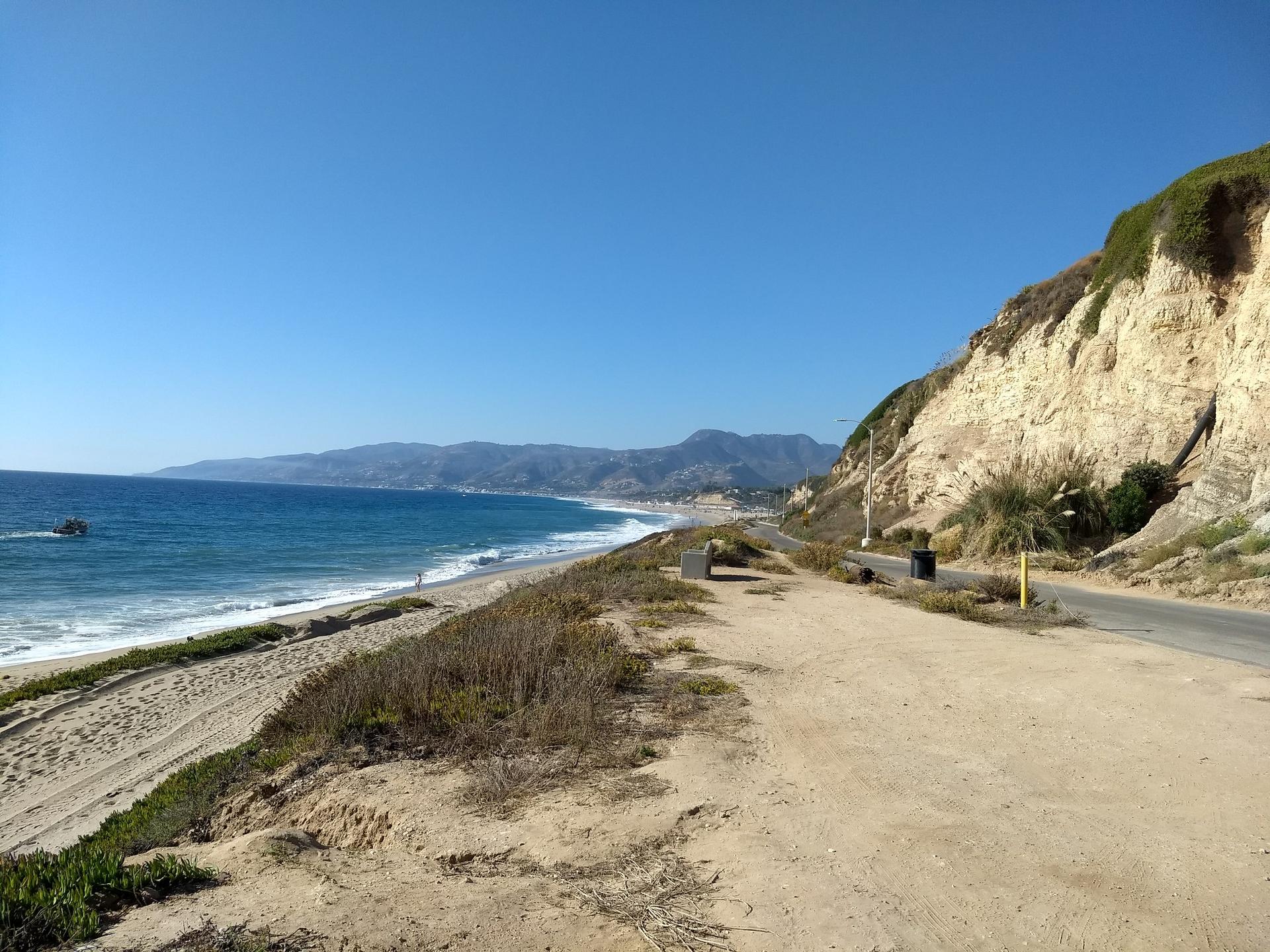 Sunny beach with ocean waves, distant mountains, a boat, and a road by a cliff.
