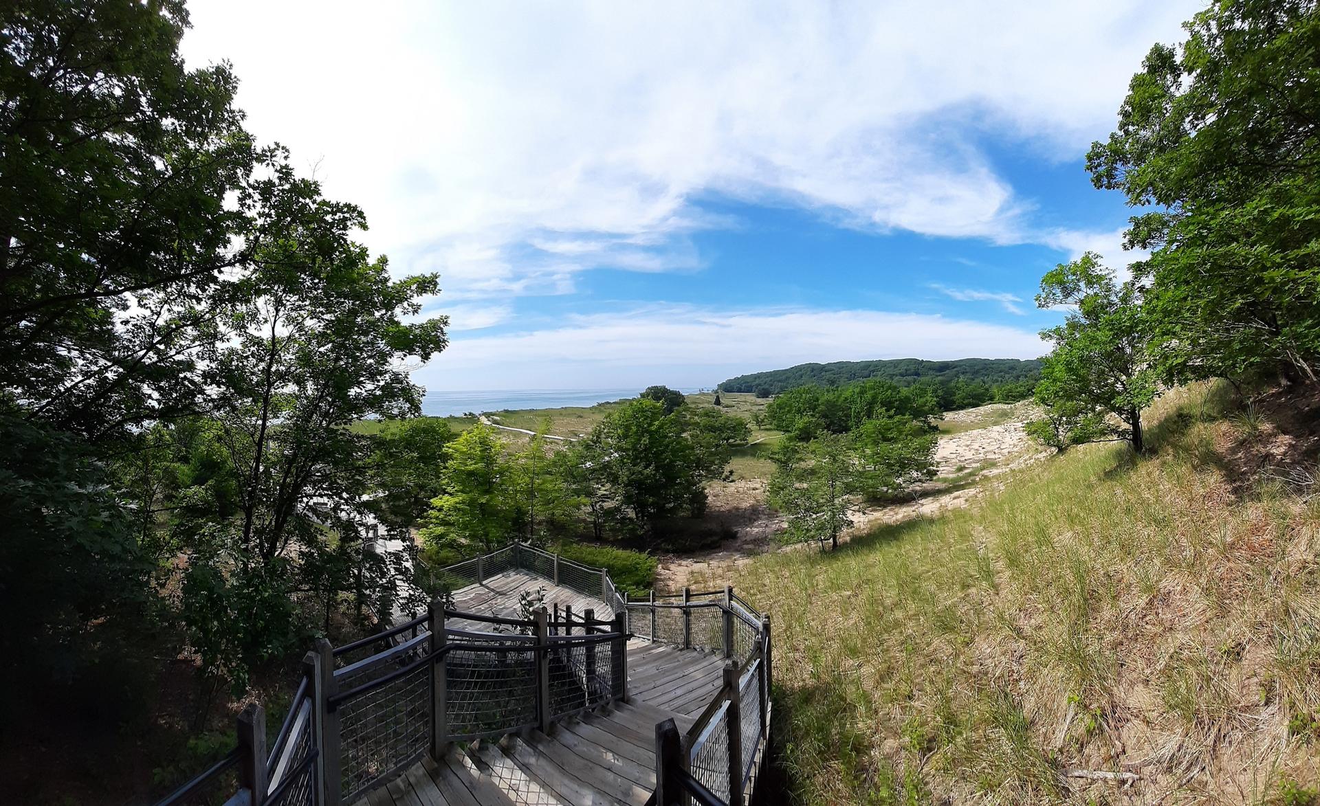 Panoramic view from wooden stairs overlooking green landscape and distant water under blue sky.