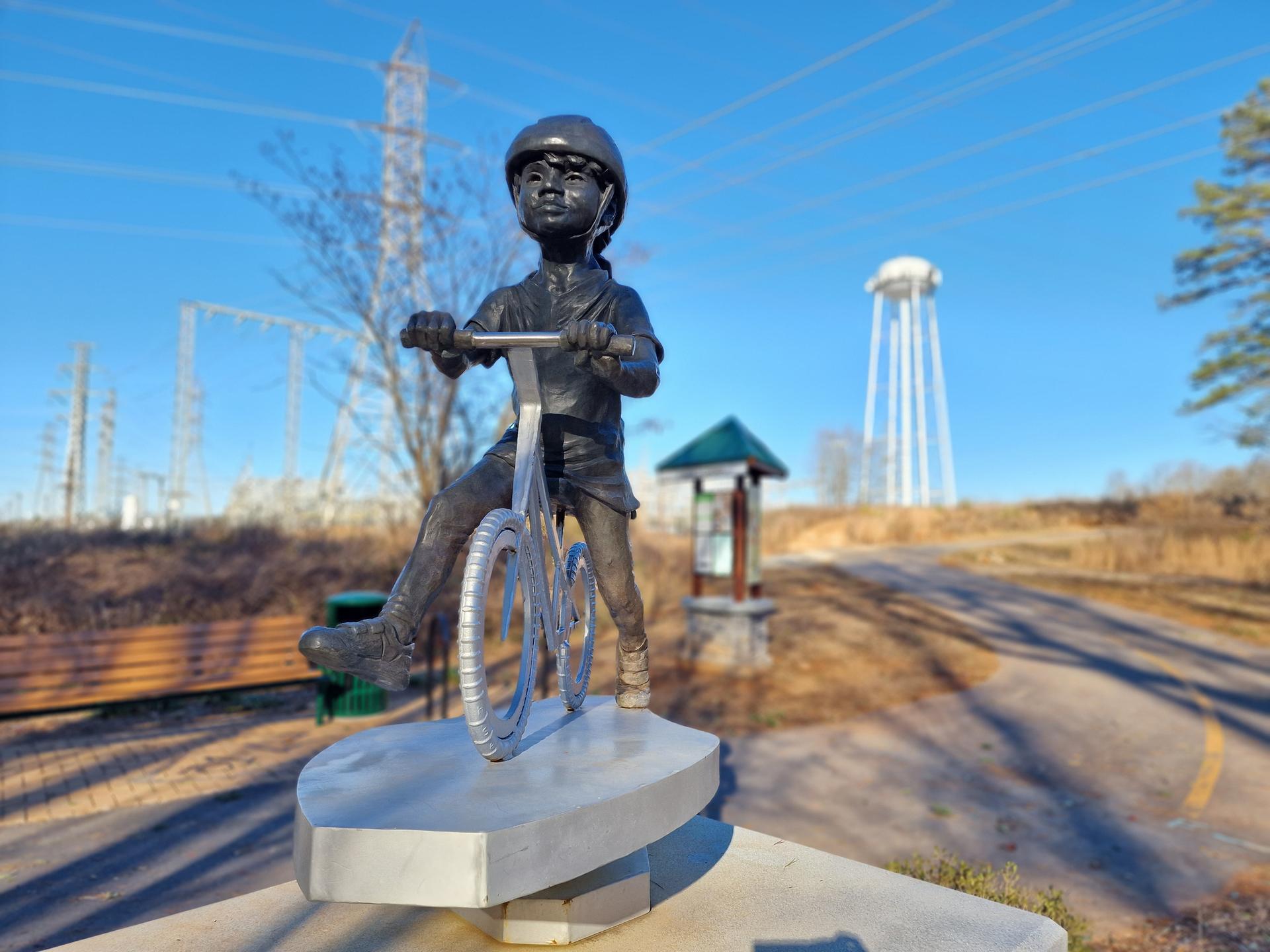 Bronze statue of a child on a bicycle on a sunny outdoor path.