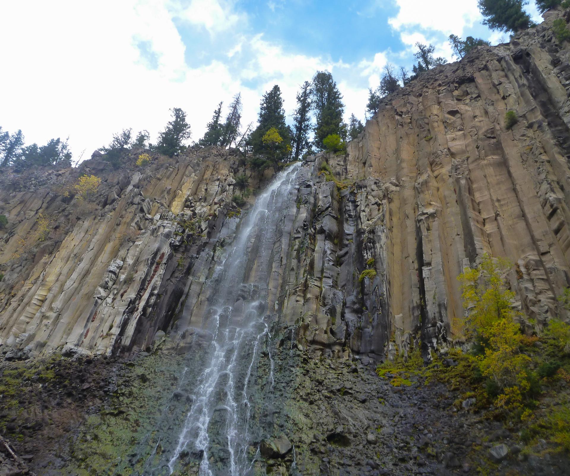 Wide waterfall flows over a textured columnar rock cliff under a blue sky.