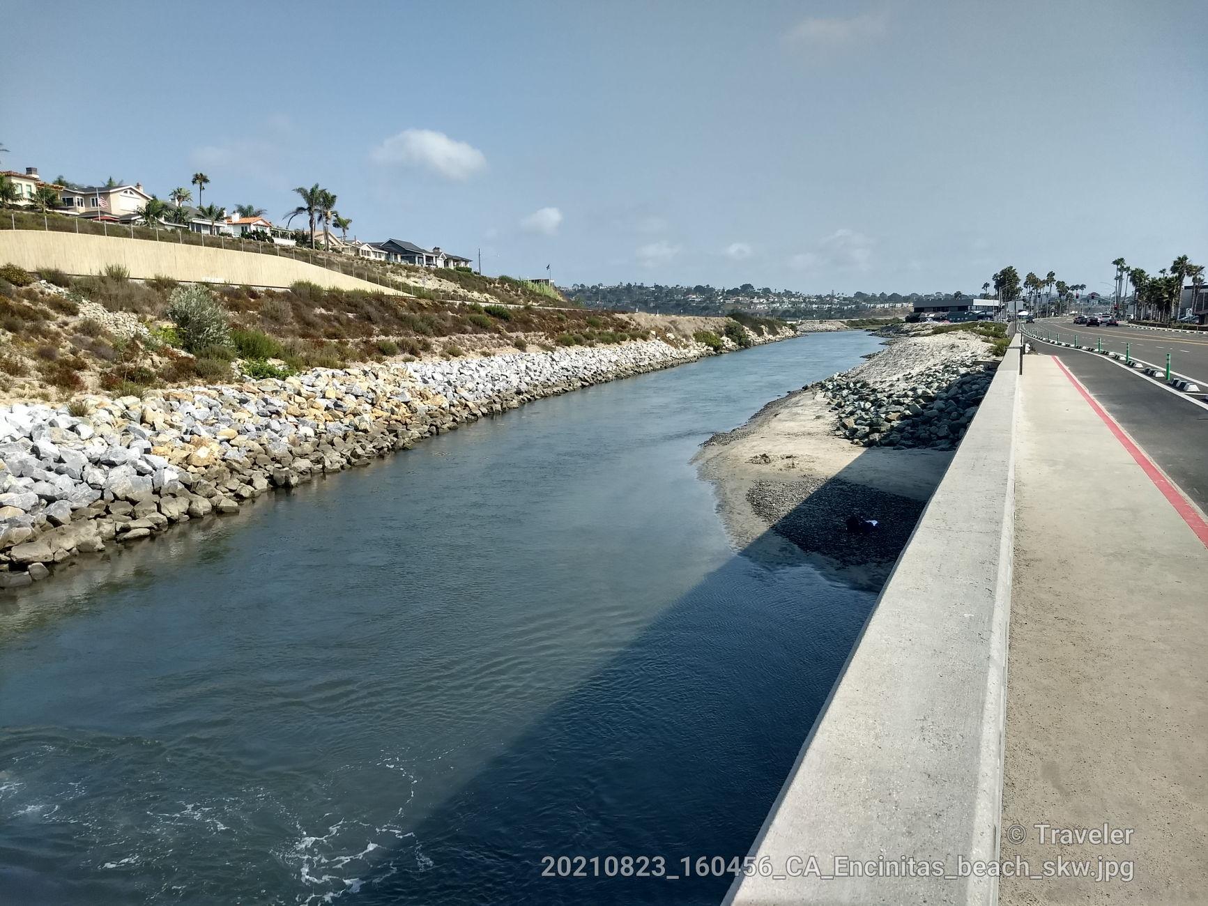 A narrow river flows between rocky banks and a road under a clear sky.