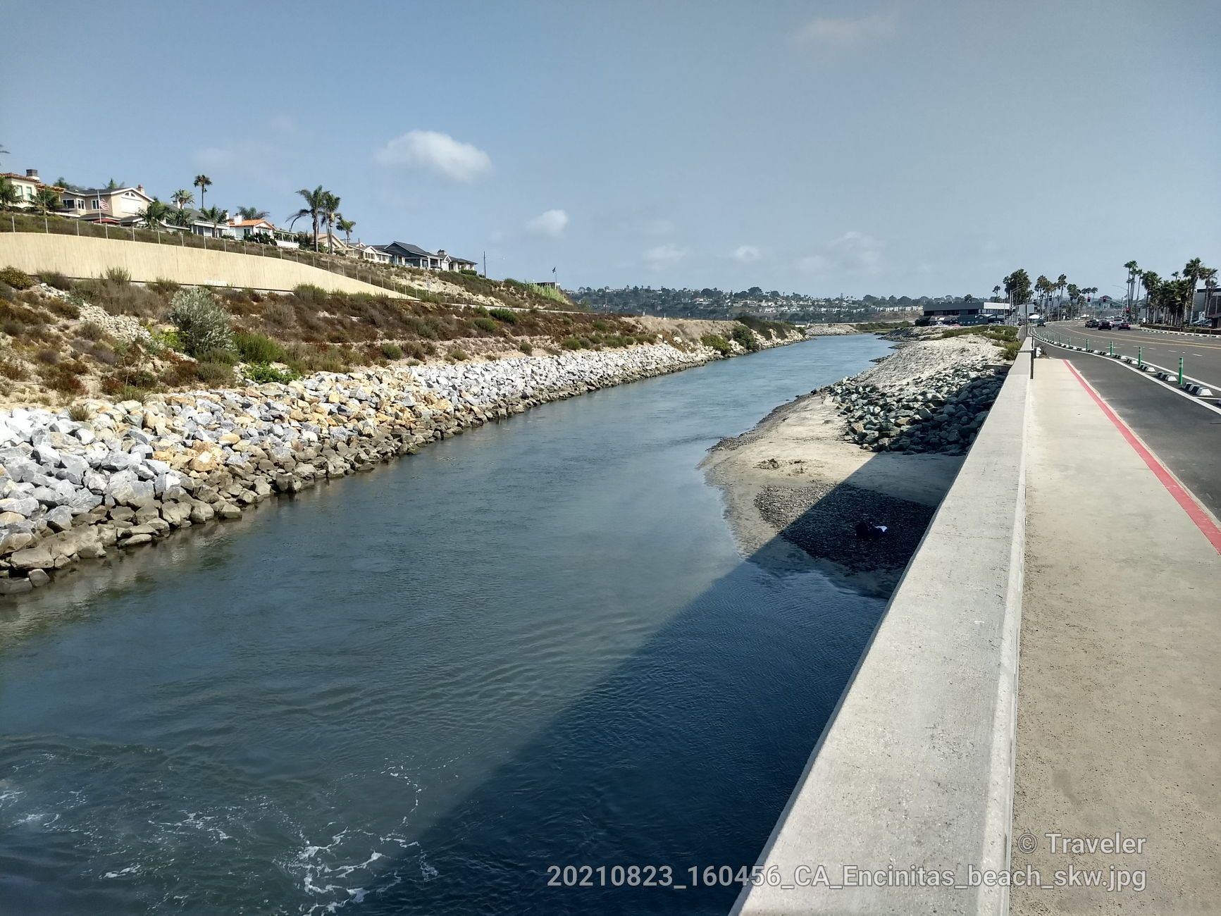 A narrow river flows between rocky banks and a road under a clear sky. - Cardiff State Beach – San Elijo Lagoon Estuary Canal loop from Solana Beach