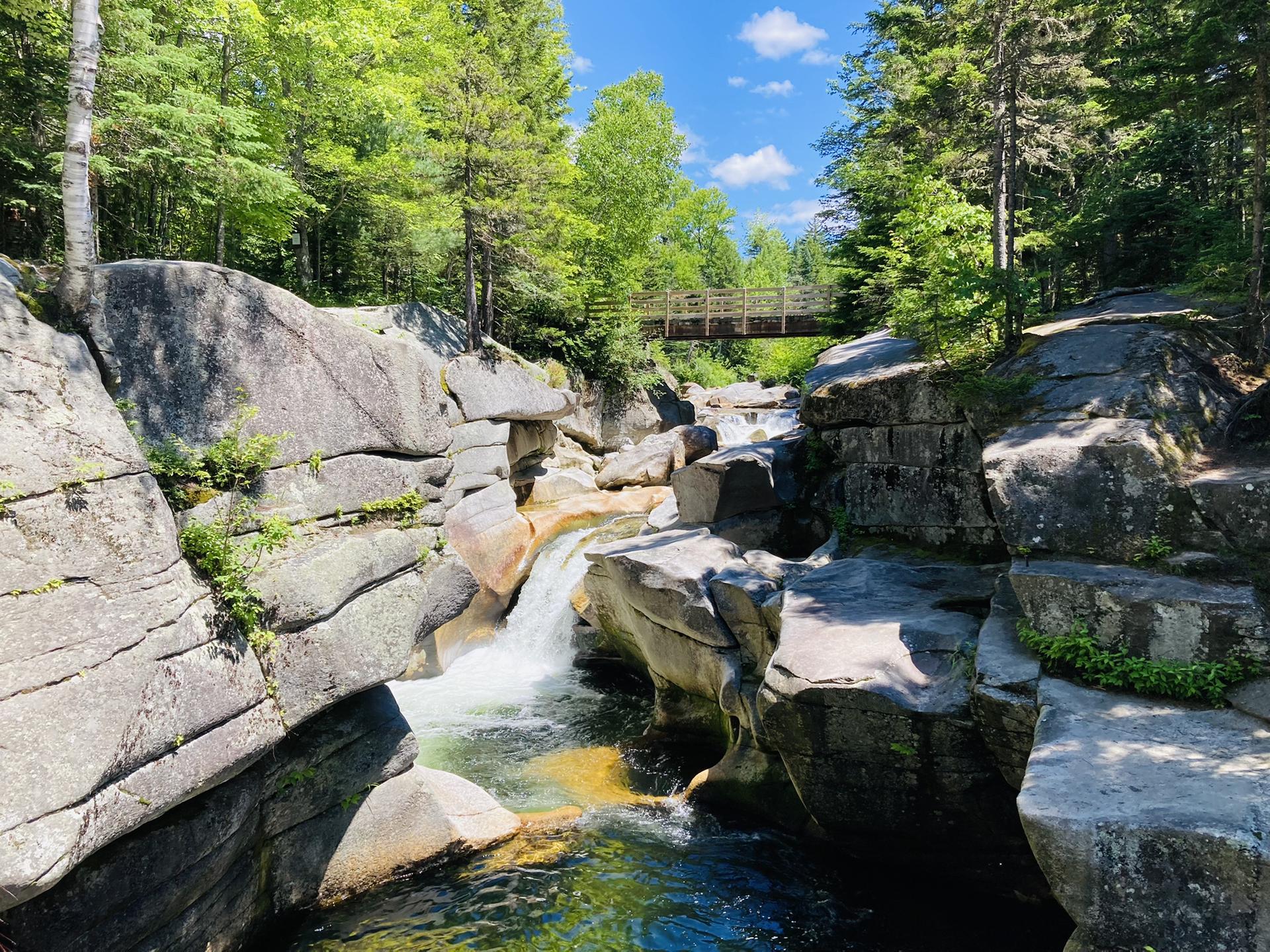 Sunny view of a clear river flowing over rocky waterfalls with a wooden bridge.
