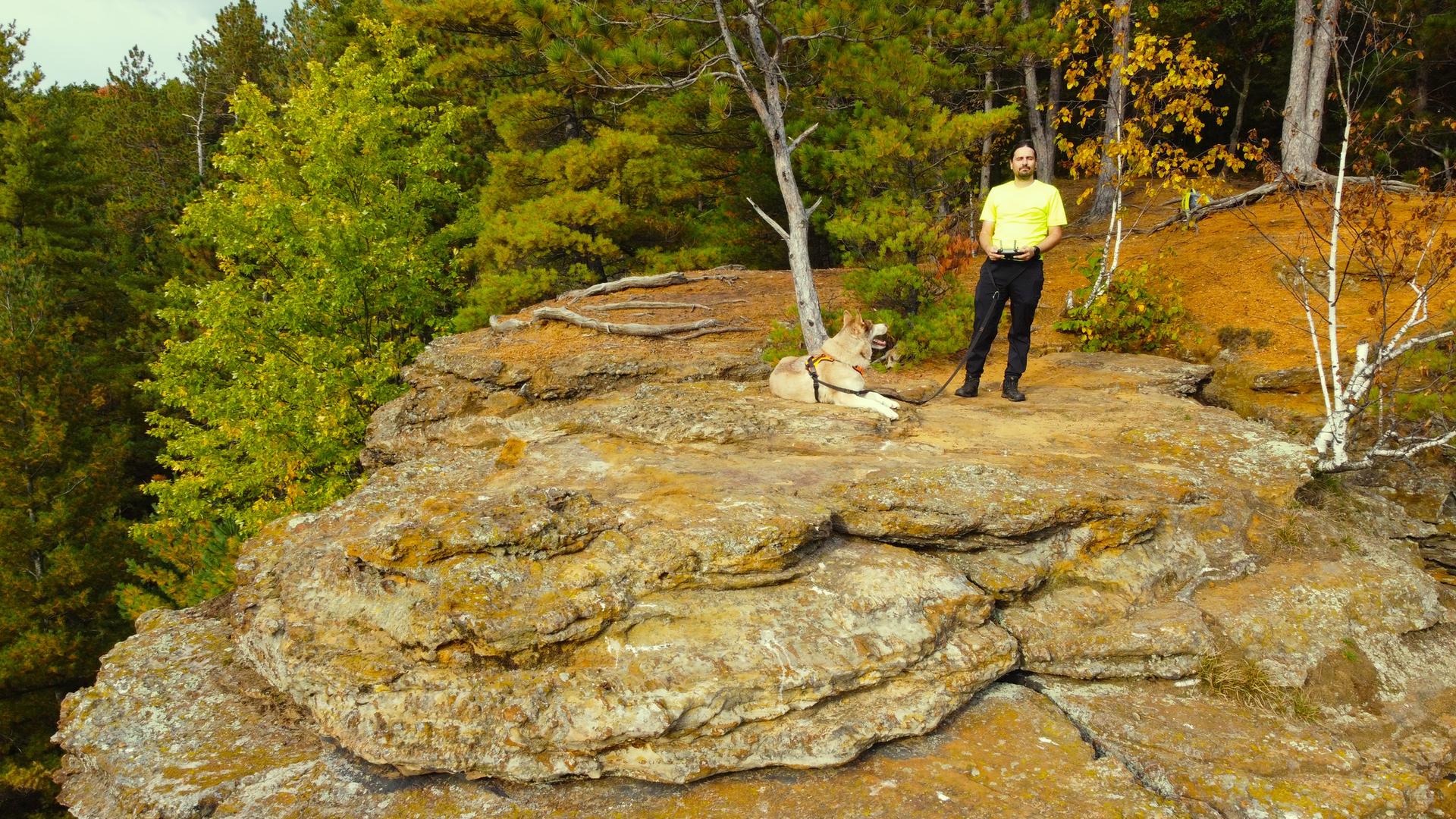 A man and his dog rest on a large rocky outcrop overlooking a forest.