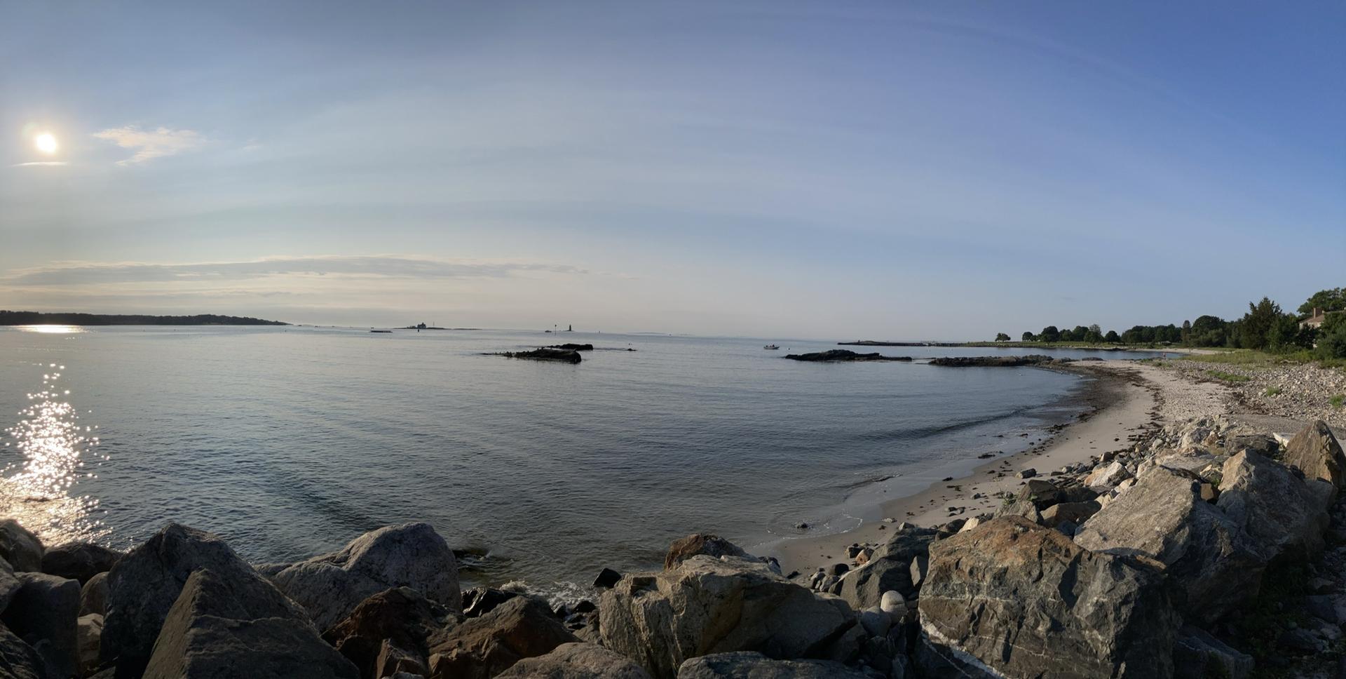 Panoramic view of a rocky beach and calm ocean under a bright, sunny sky.