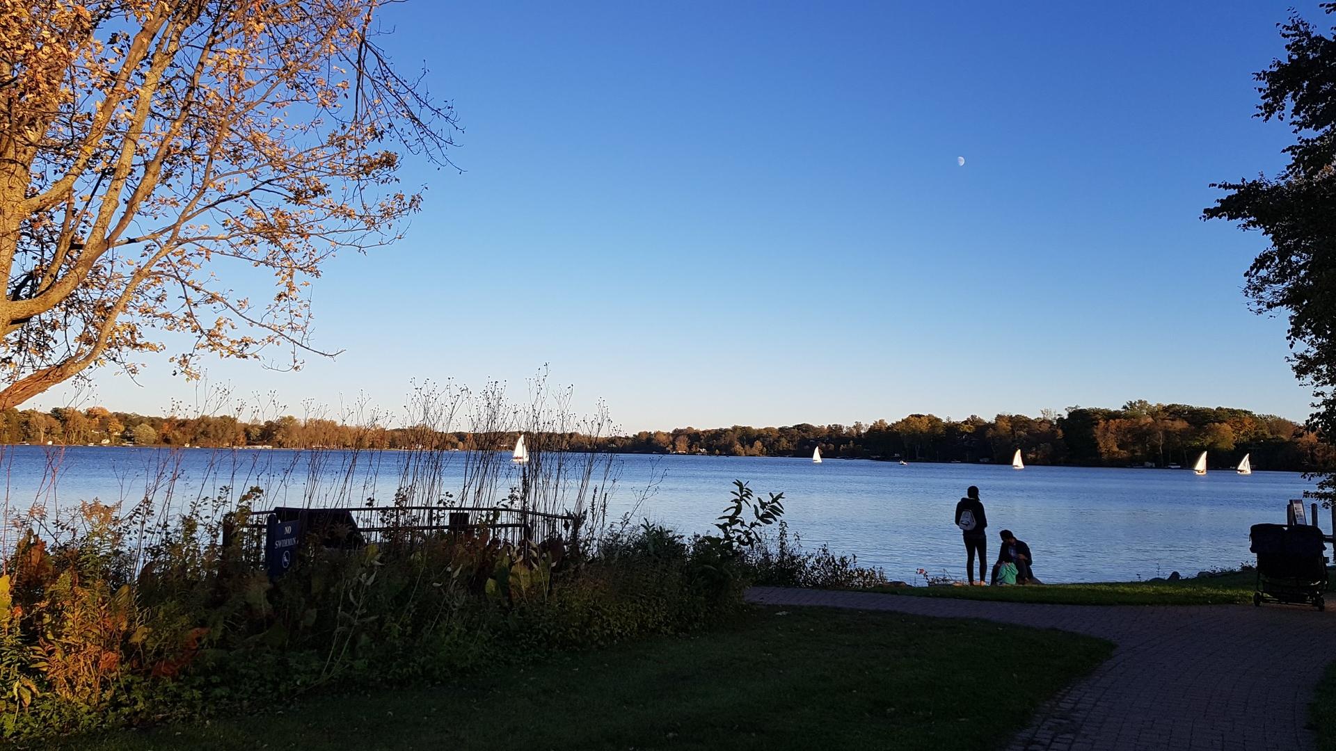 Two people stand by a lake with sailboats under a clear evening sky.