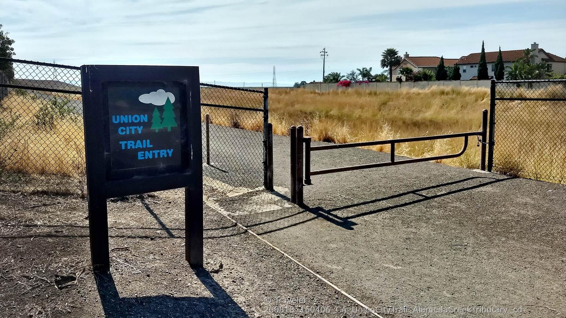 Trail entry sign next to a fence and gate on a sunny day.