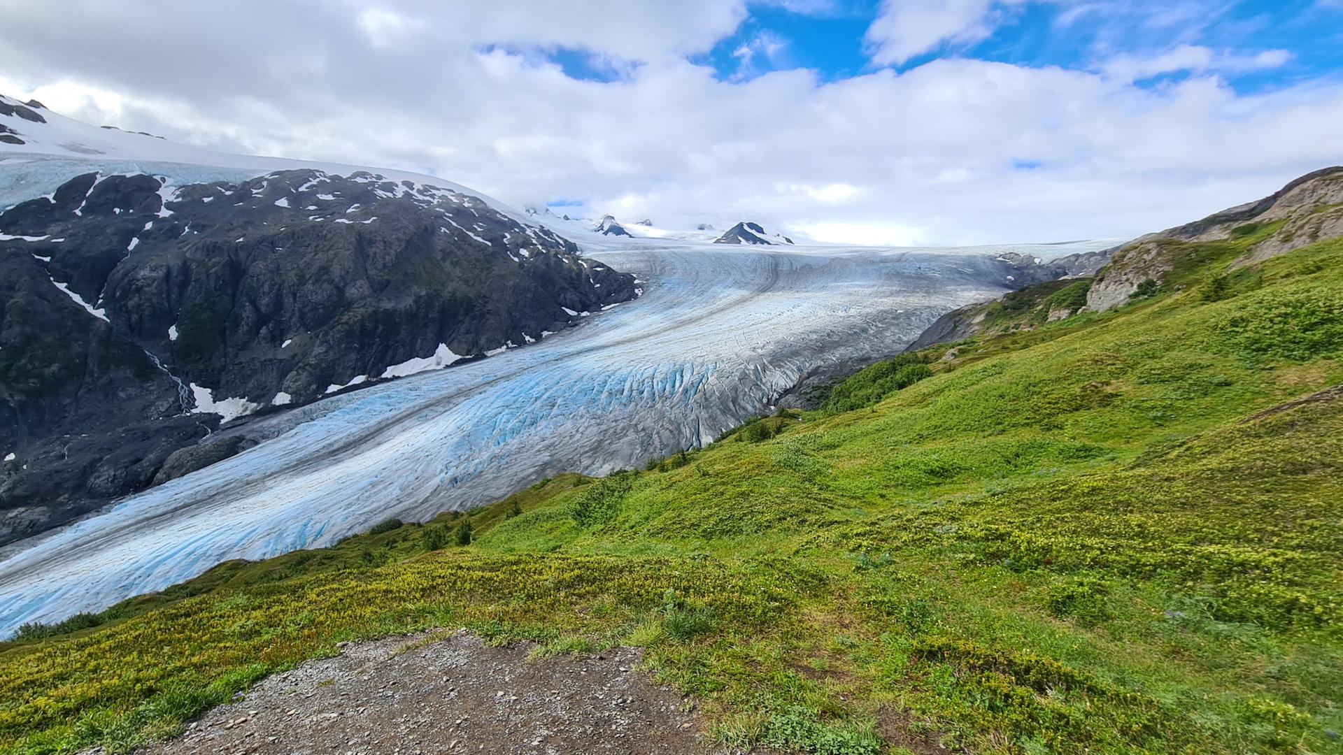 Vast blue glacier flows between dark mountains and vibrant green hills under cloudy sky.
