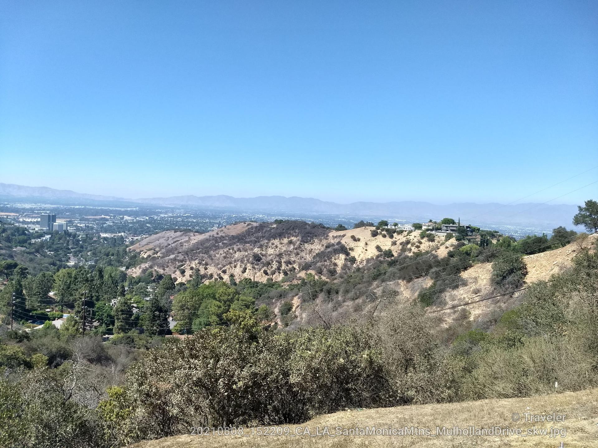 Expansive view of dry hills, green trees, and a distant city under clear skies.