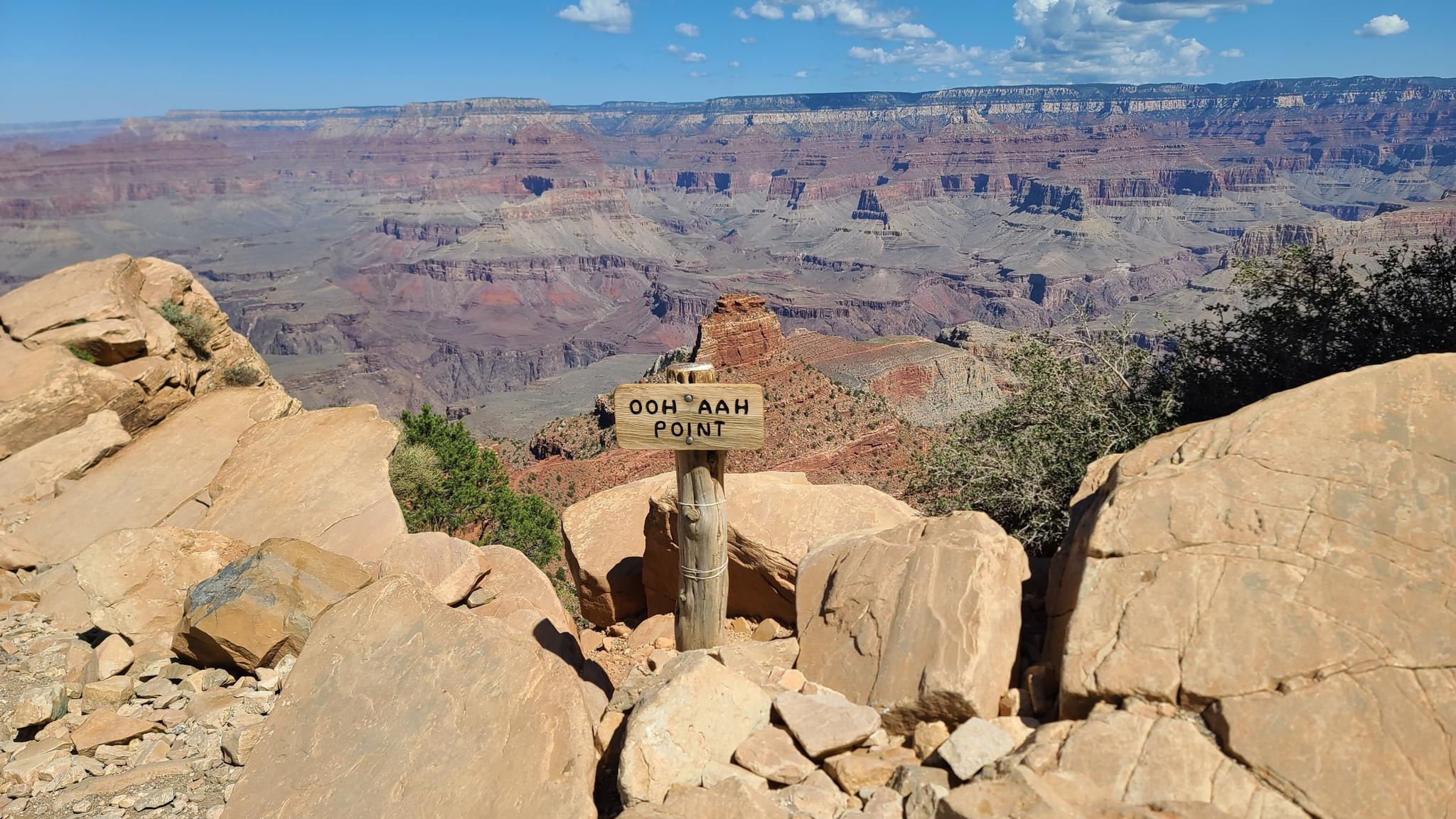 Wooden sign on a rocky outcrop overlooking a vast, sunlit canyon.