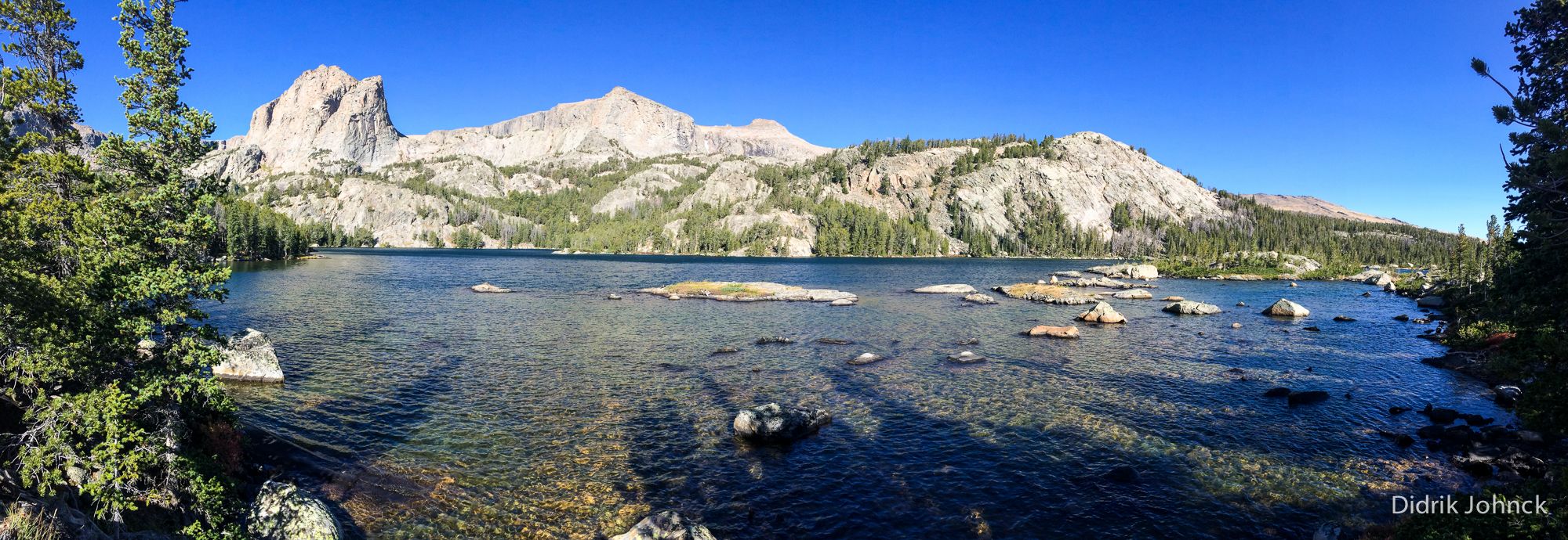 Sunny panoramic view of a clear lake with mountains and trees. - Glacier Trail